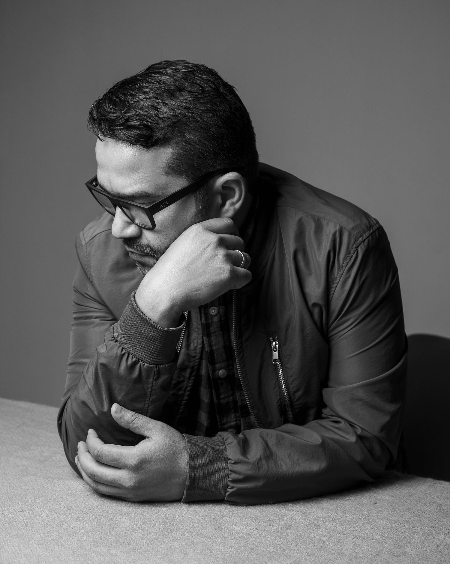 Man with glasses in black and white portrait, thoughtfully resting chin on hand while looking down.