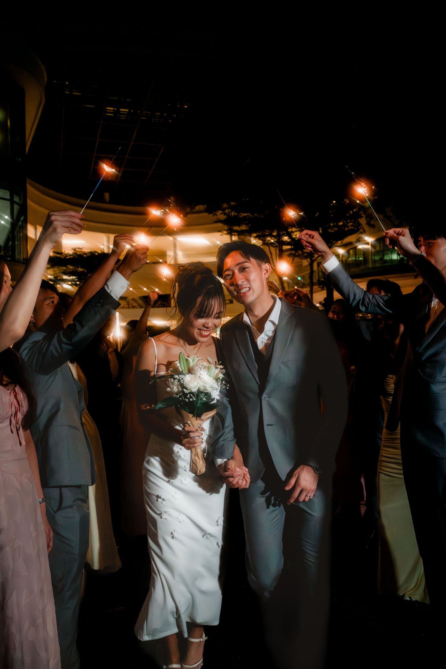 Newlyweds walk through sparkler tunnel at night wedding reception, bride holding white bouquet in elegant white dress.