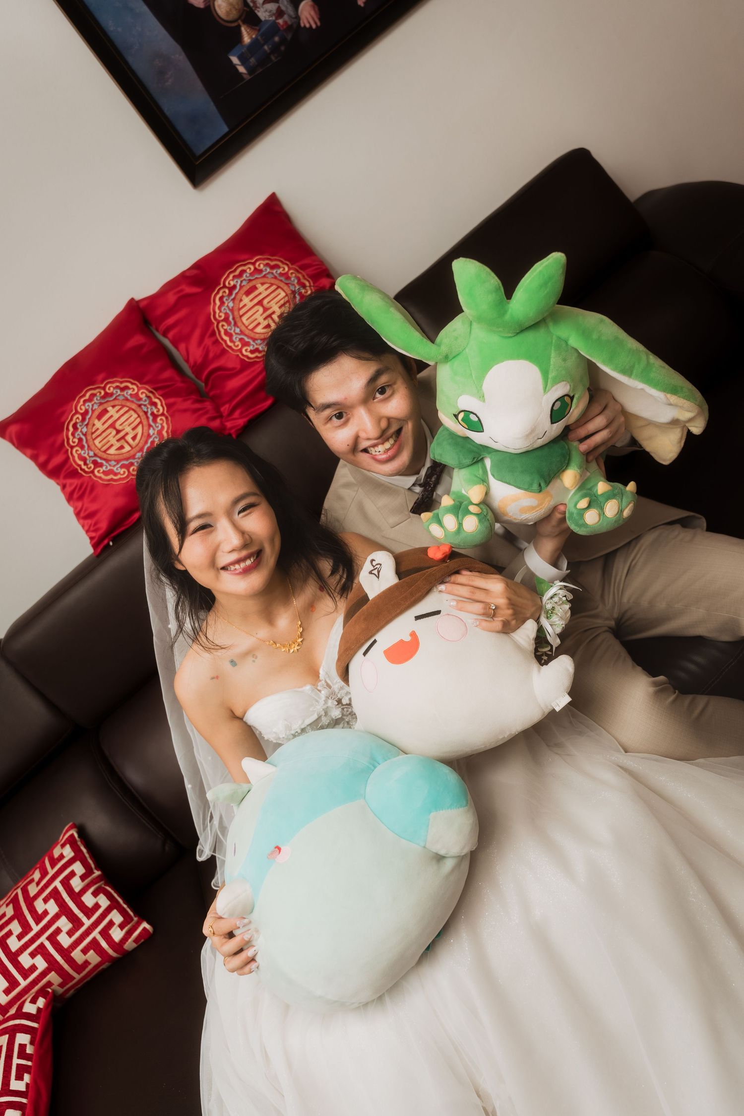 A smiling couple in wedding attire sits on a sofa holding colorful plush toys, with red decorative pillows behind them.