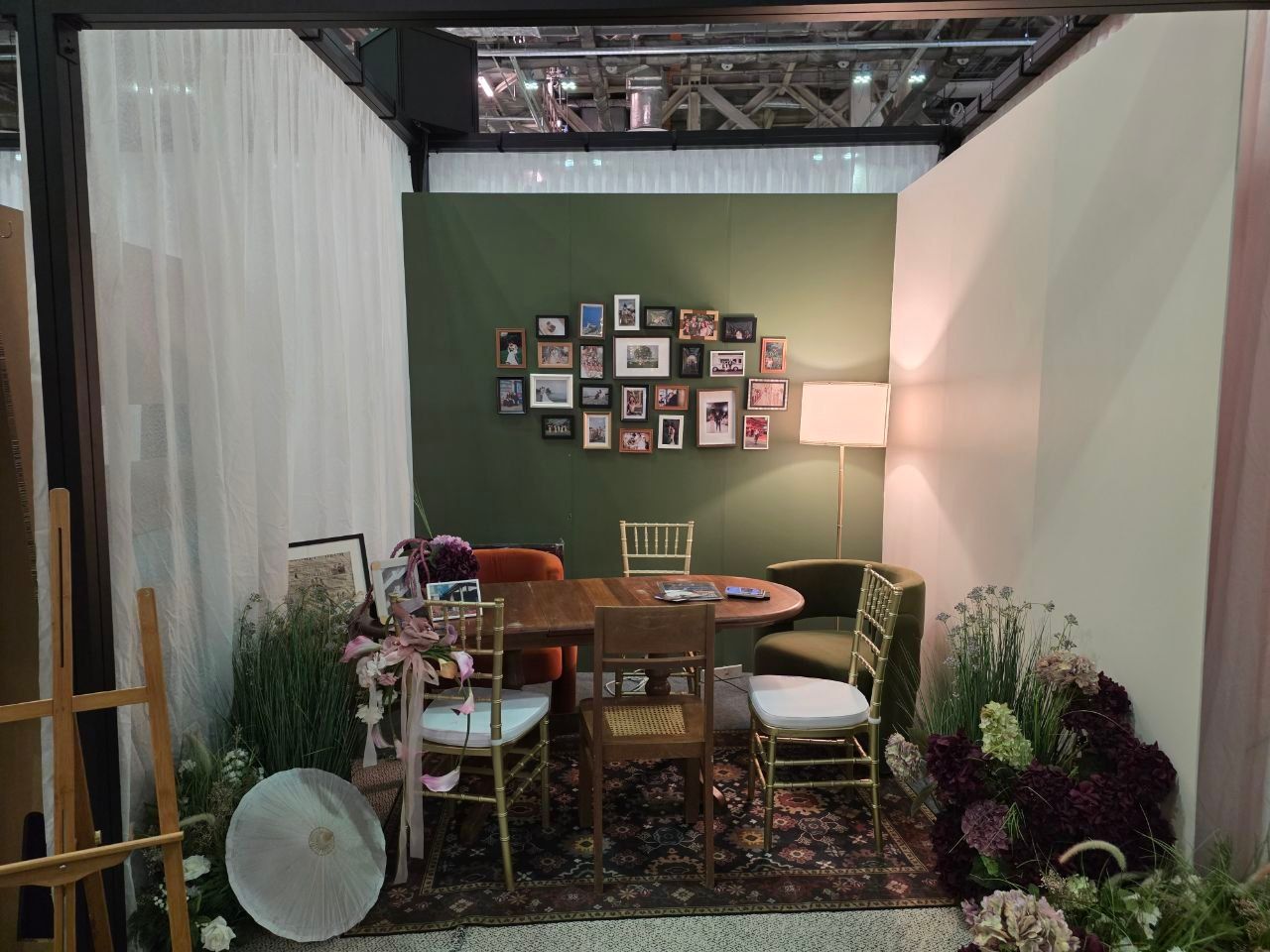 Cozy booth display with round wooden table, gold Chiavari chairs, patterned rug, green accent wall with photo gallery, and fresh florals.
