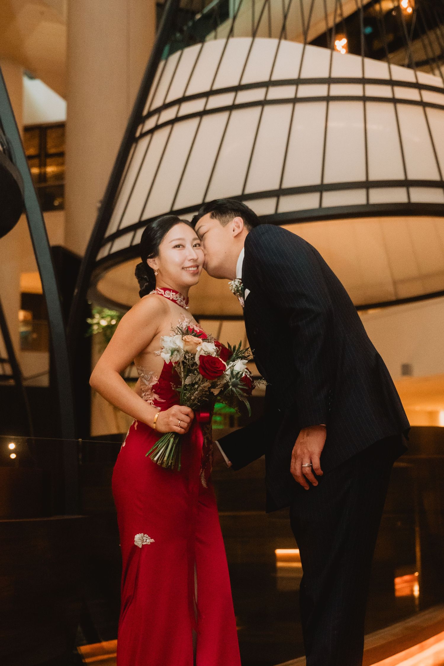 A couple in formal attire shares a romantic kiss, woman in red gown holding a bouquet inside an elegant venue.