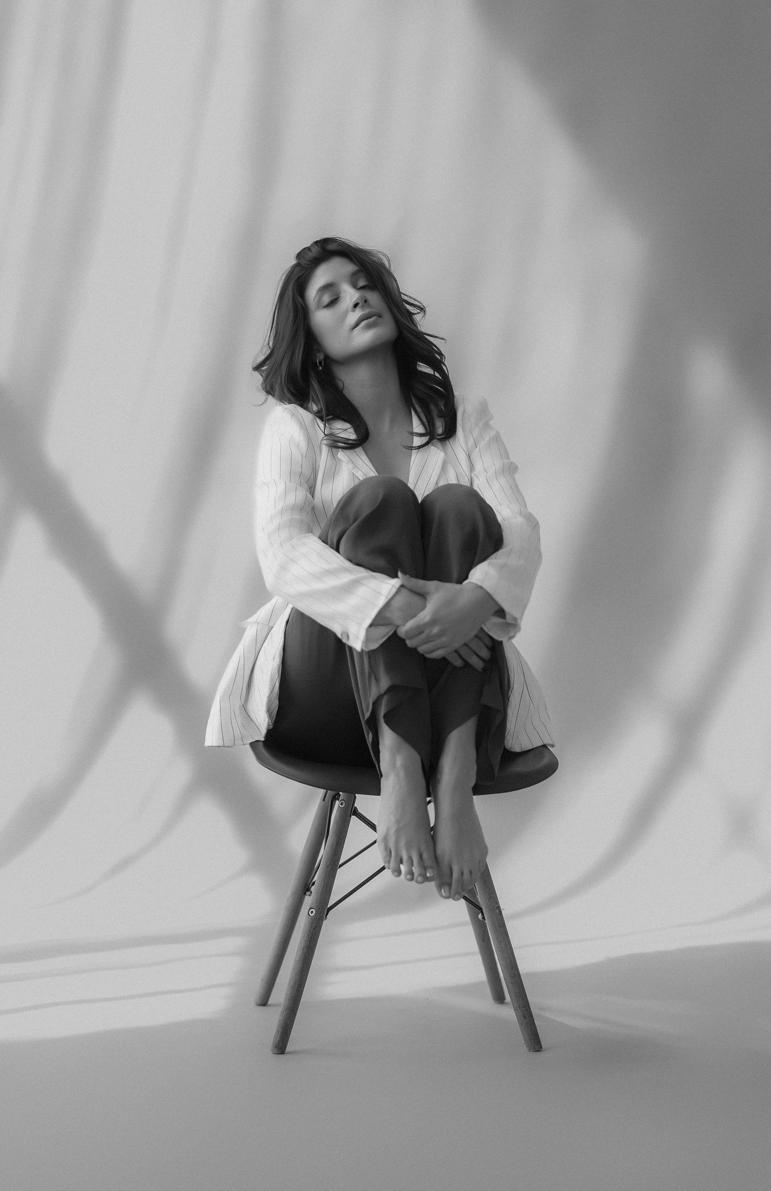Black and white artistic portrait series showing figure in white shirt seated on chair against light backdrop with shadows.