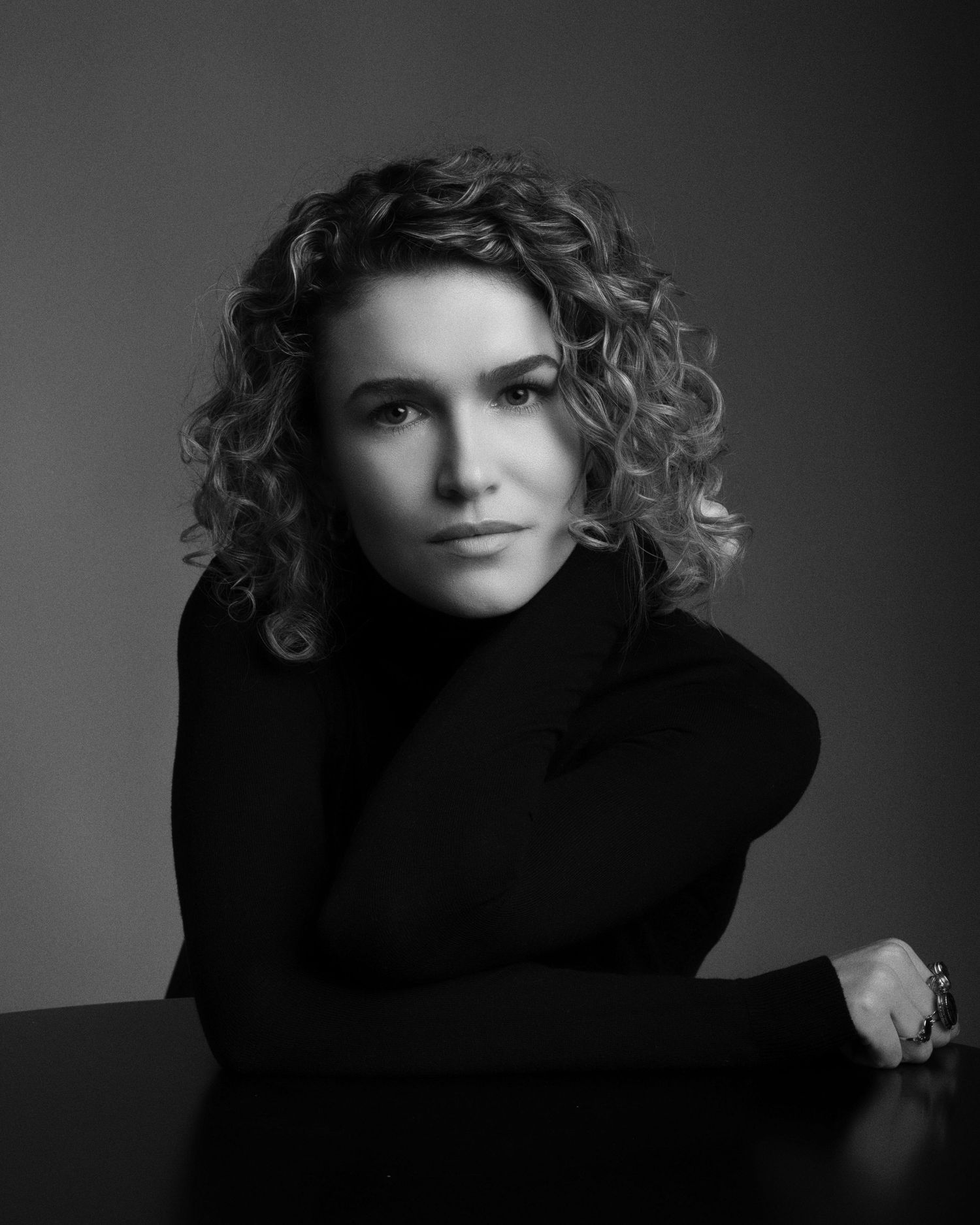 Black and white portrait of a person with curly hair wearing a black turtleneck sweater against a dark background.