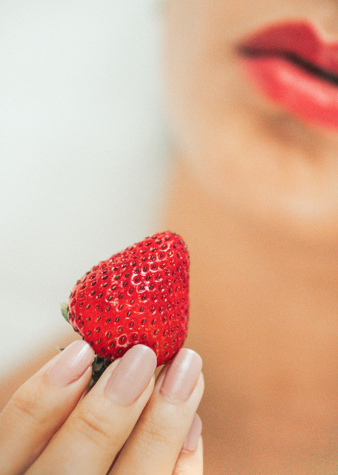 Elegant photo of perfectly ripe strawberry held by manicured fingers with red lips in soft focus.