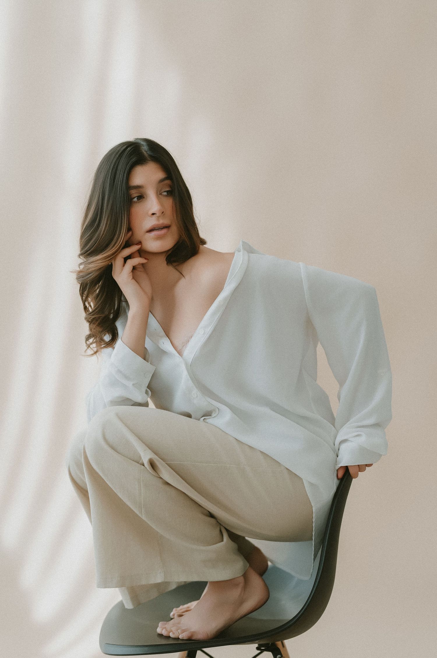 Artistic fashion portrait of person in white oversized shirt sitting in chair against light neutral background.