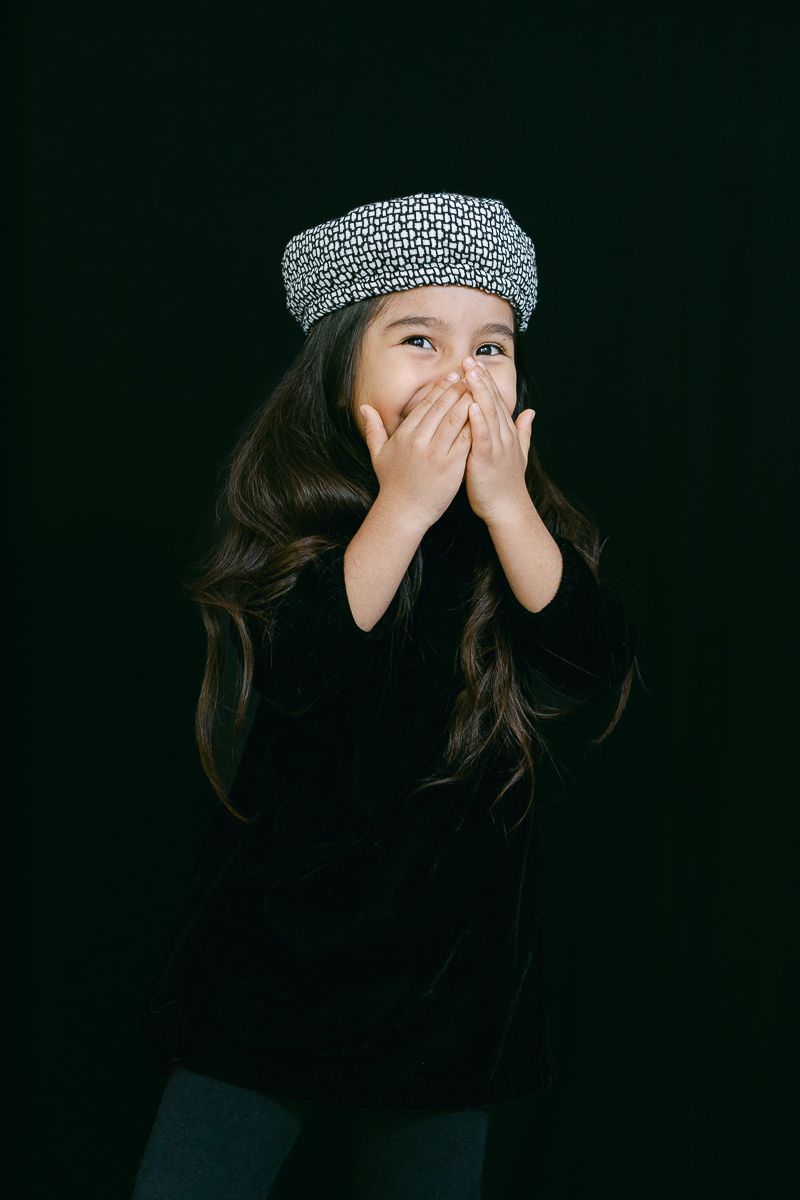 Playful portrait with hands covering mouth while wearing a patterned beret and black top.