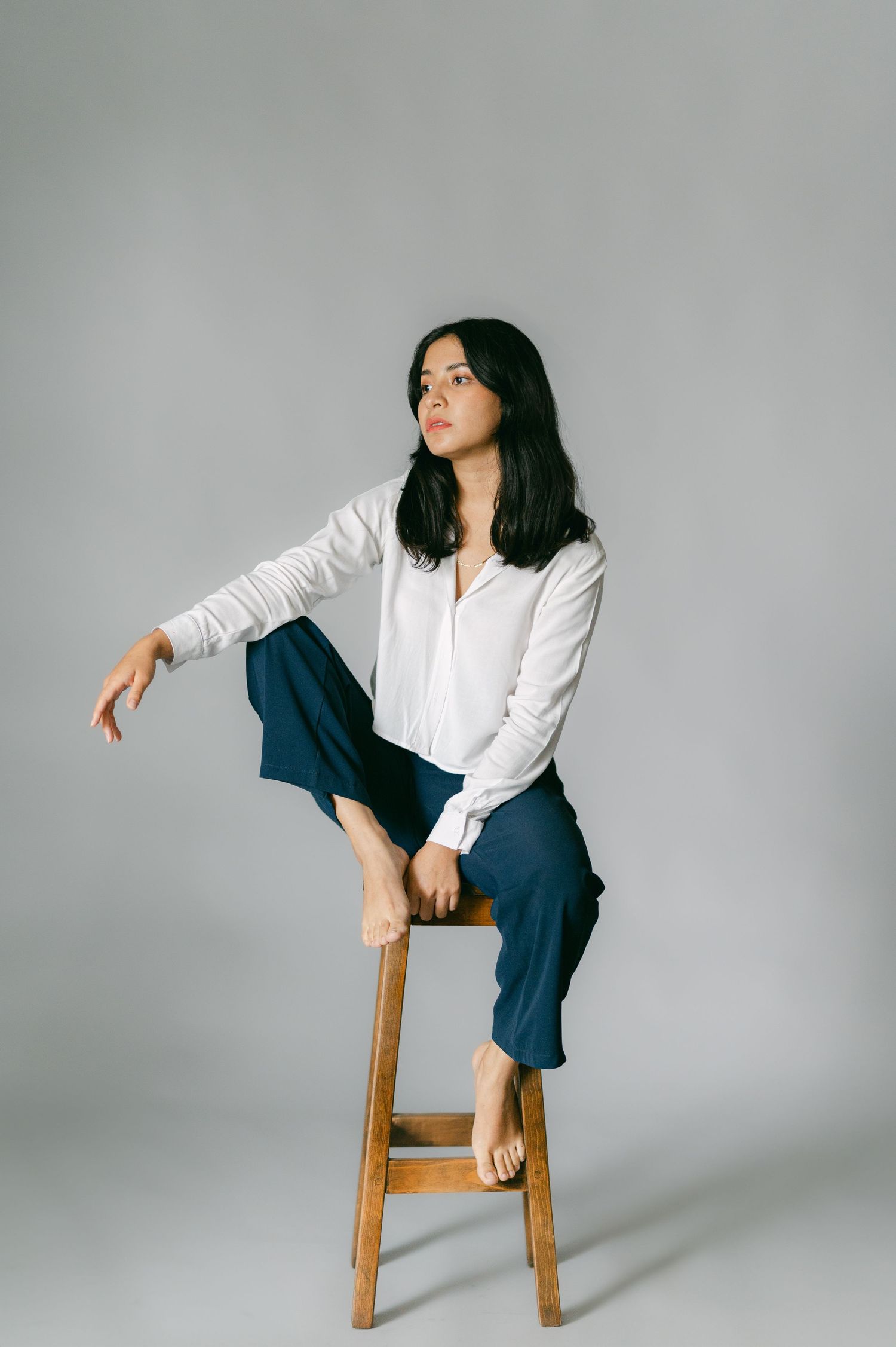 Person in white blouse and blue pants sitting casually on wooden stool in studio setting.