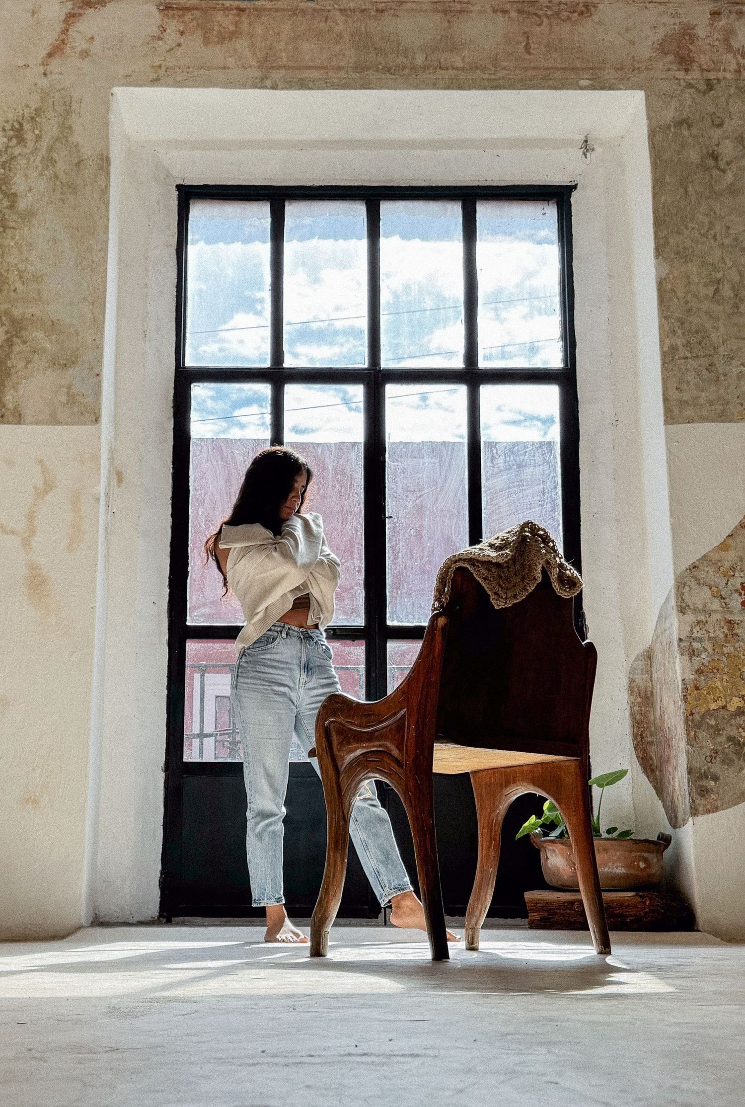 Woman standing by vintage window with antique wooden chair in rustic room.