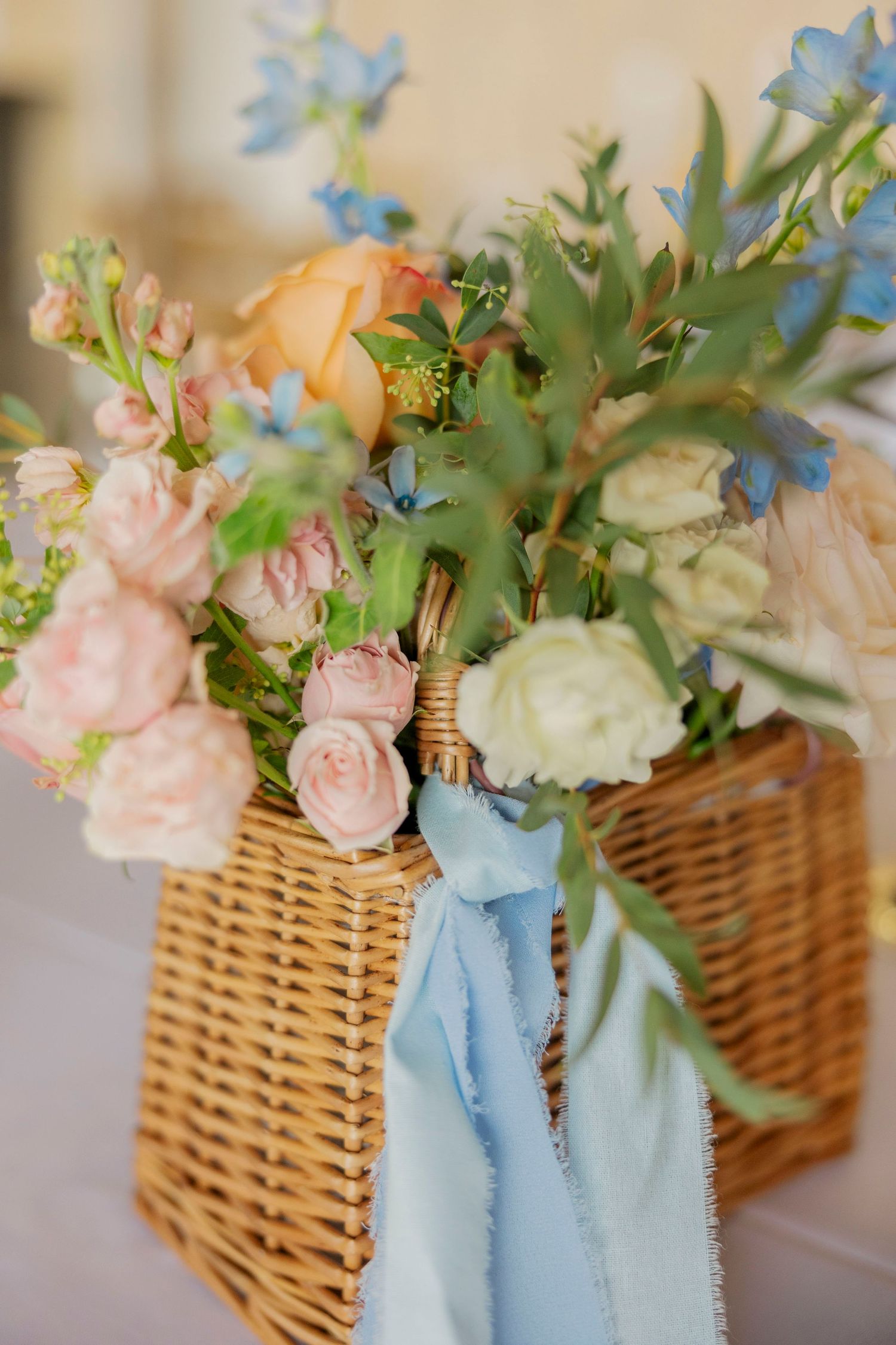 Wicker basket arrangement with pink roses and blue flowers tied with light blue ribbon.