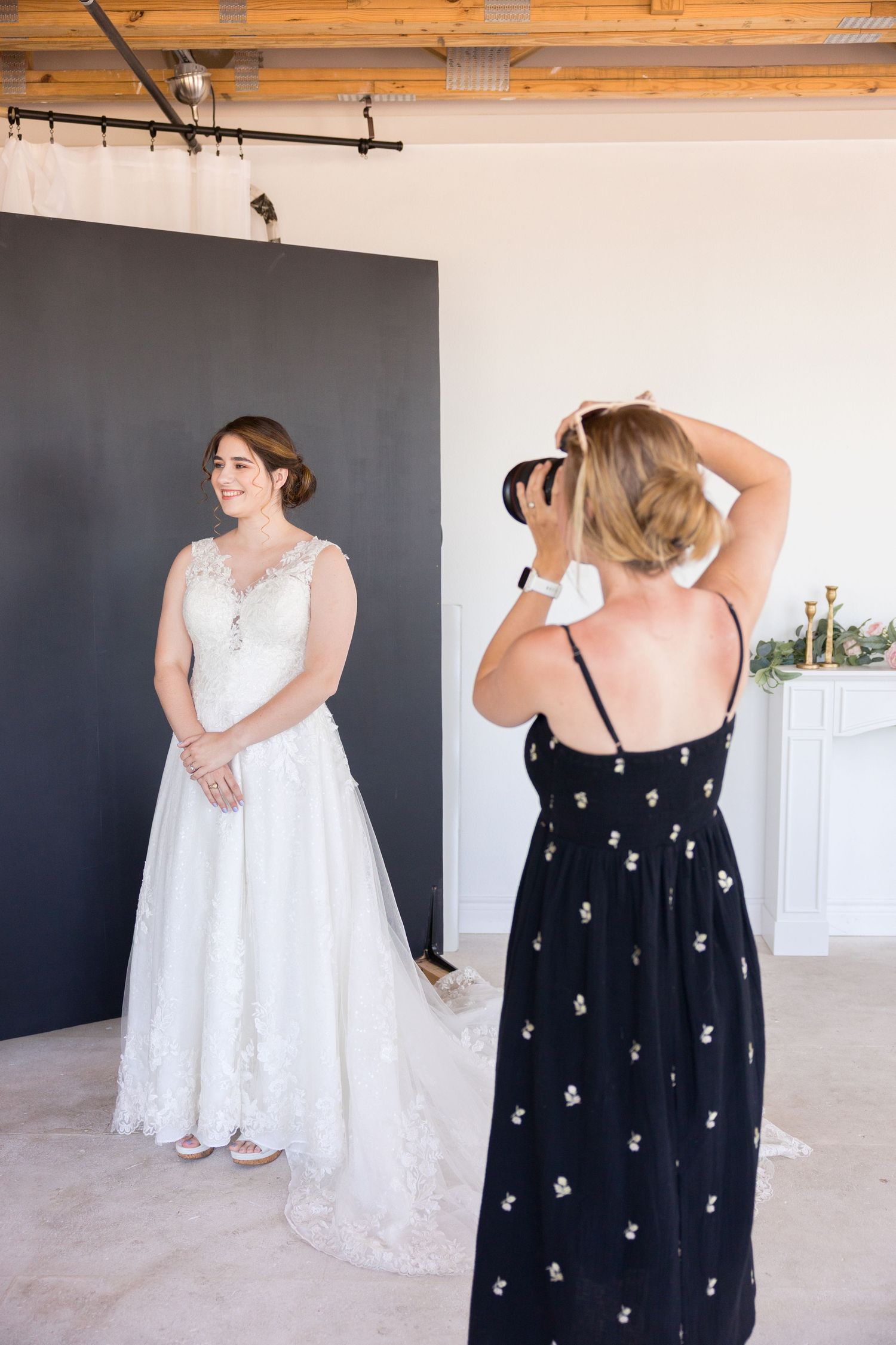 A photographer in a black floral dress captures wedding photos in a modern indoor setting with rustic wood beams.