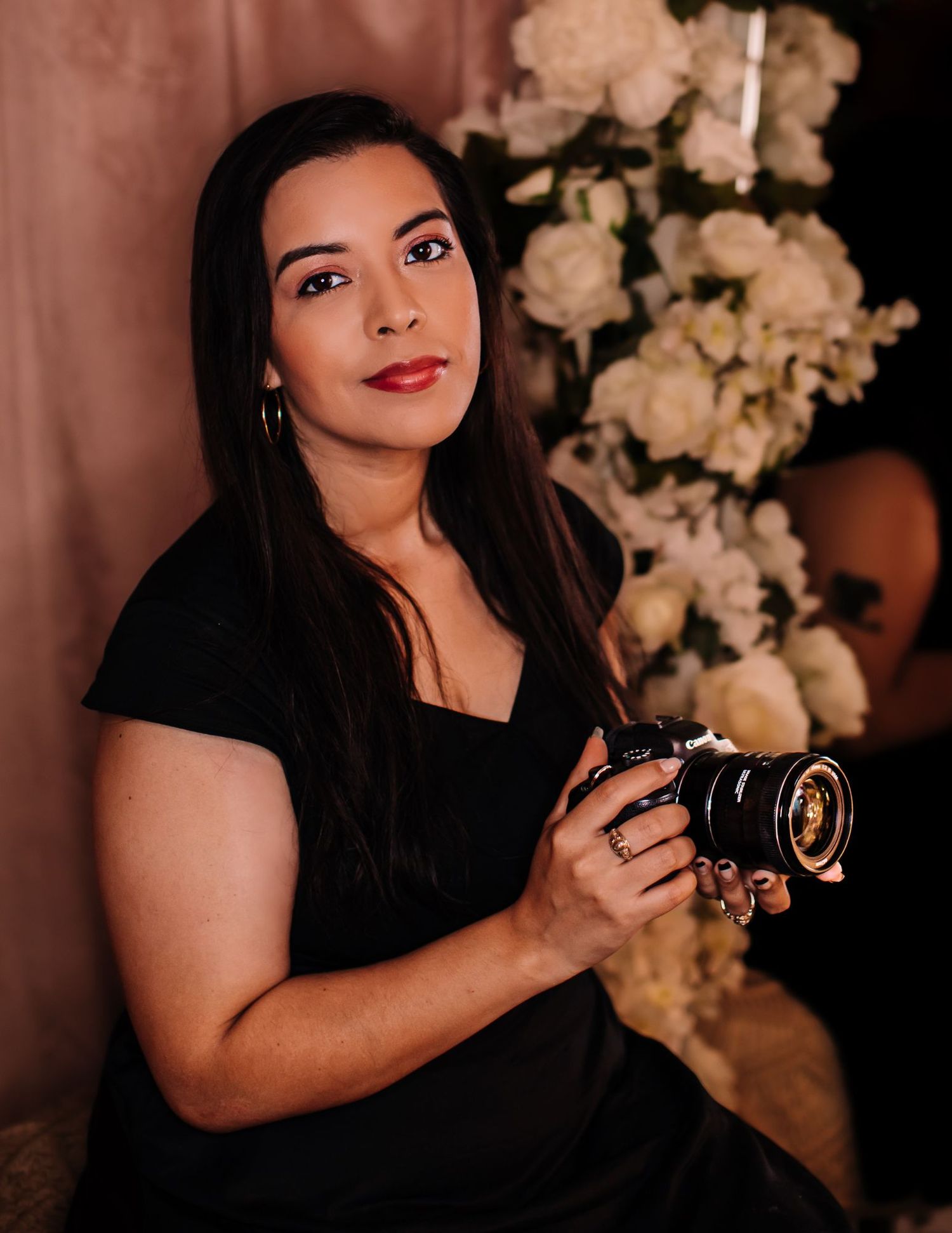 Woman in black dress holding a professional camera, posed elegantly in front of white floral arrangement.