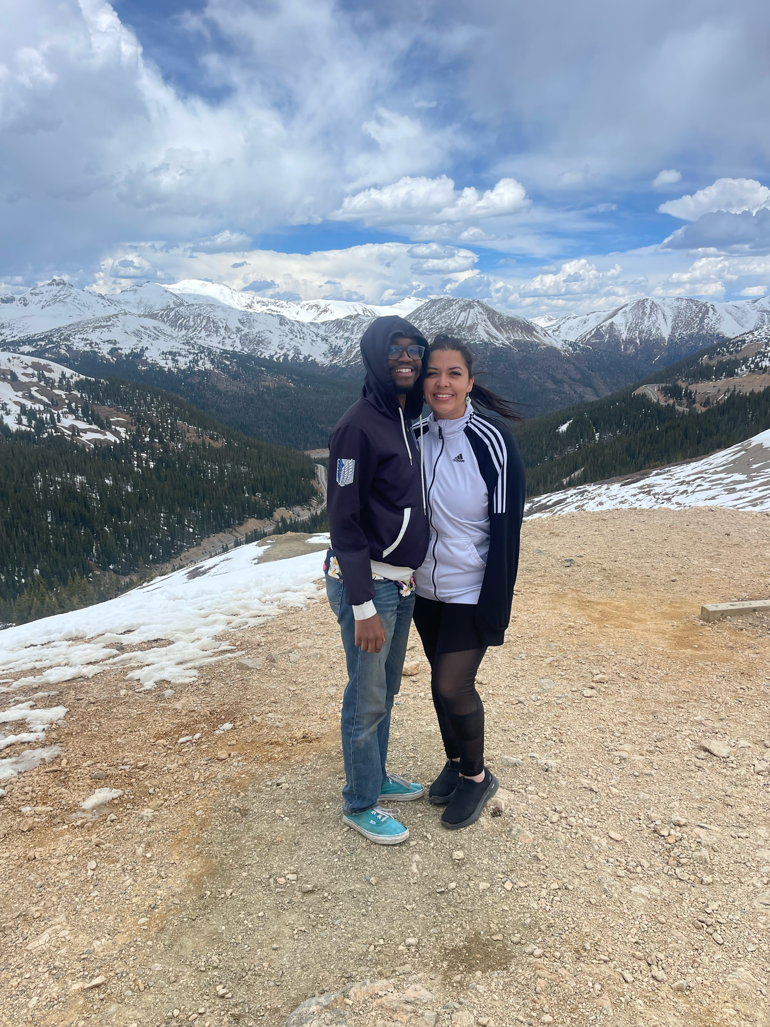 Couple posing together on a snowy mountain trail with stunning Rocky Mountain peaks and evergreen forests in the background.