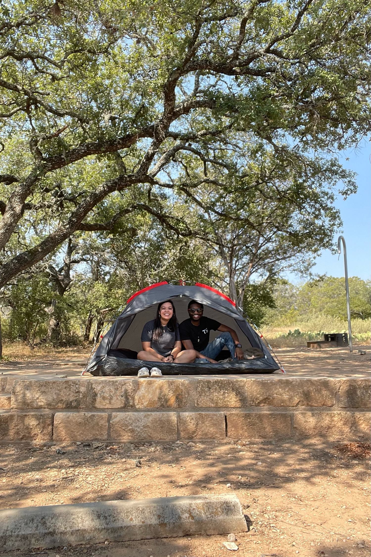 Two people sit smiling inside a dome tent at a shaded campsite beneath large oak trees on a sunny day.