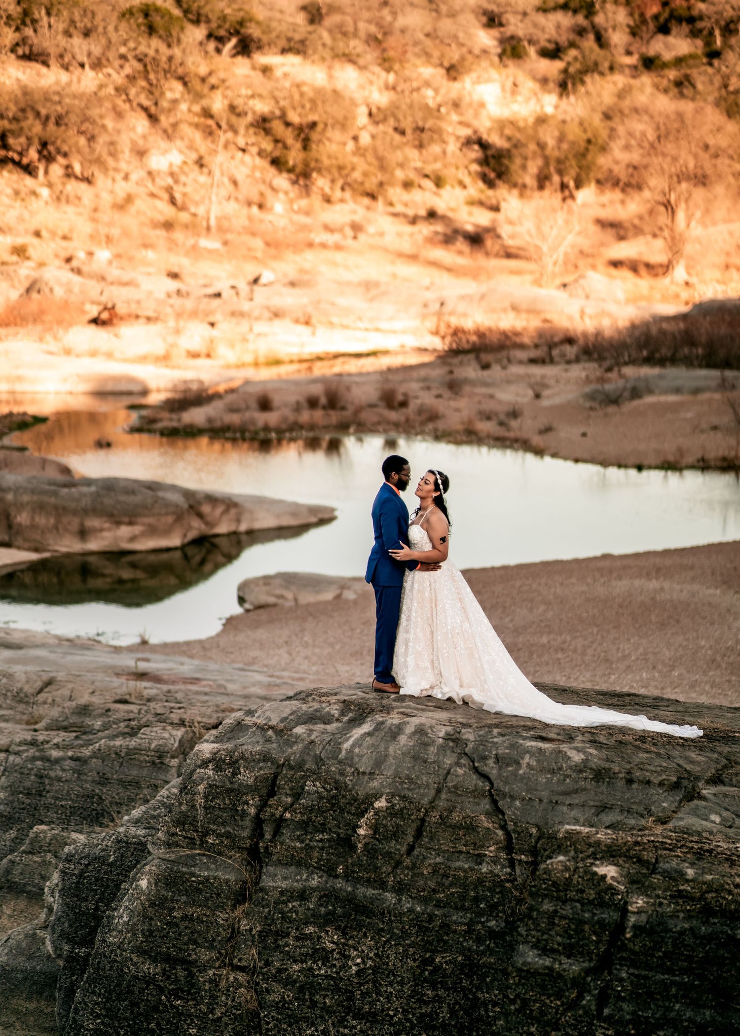 Bride and groom stand on rocky riverbank at golden hour, wearing wedding gown and navy suit, with scenic water backdrop.