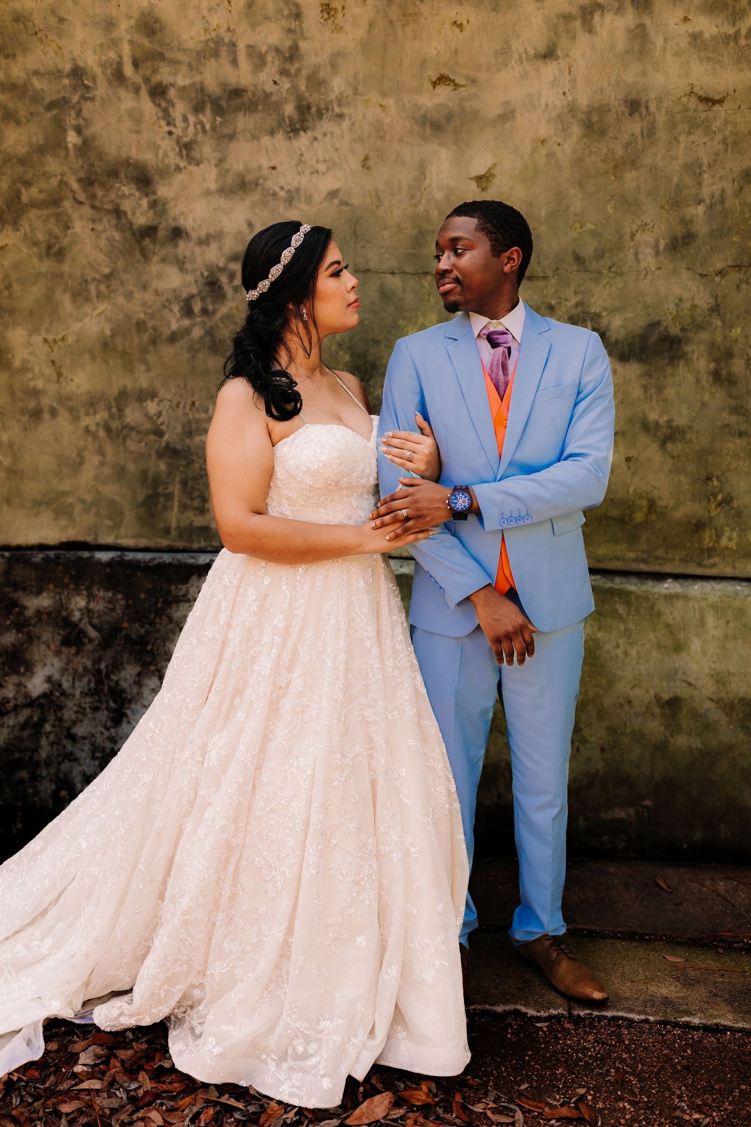 Bride in white lace gown and groom in light blue suit with orange tie share a romantic moment outdoors.
