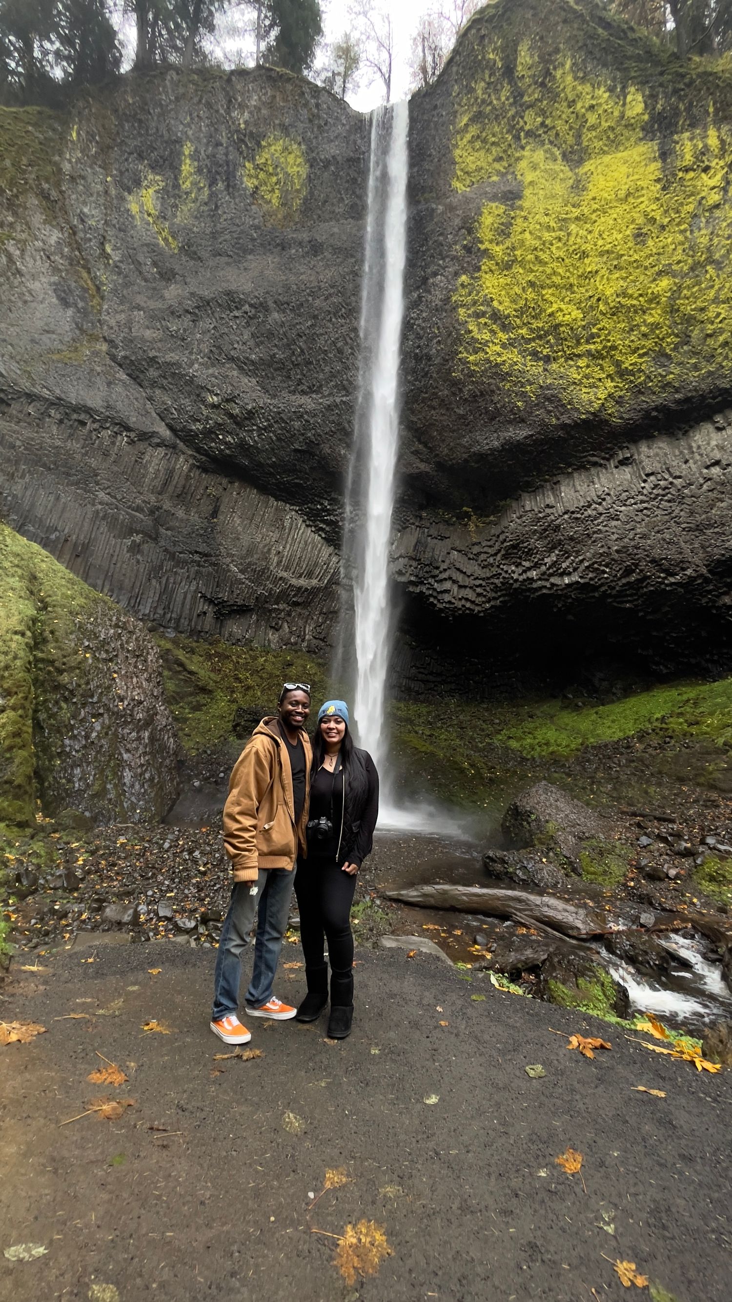 Couple posing in front of a tall waterfall cascading down mossy basalt cliffs surrounded by autumn foliage.
