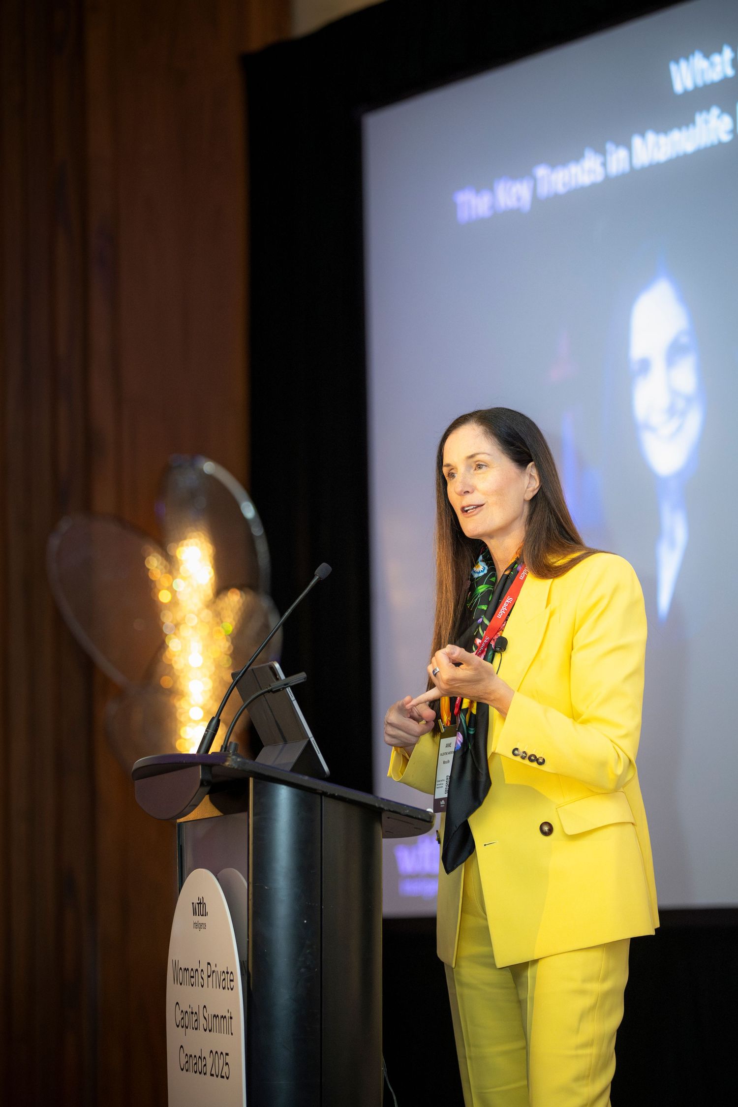 A presenter in a bright yellow blazer speaks at a podium during a professional conference presentation.
