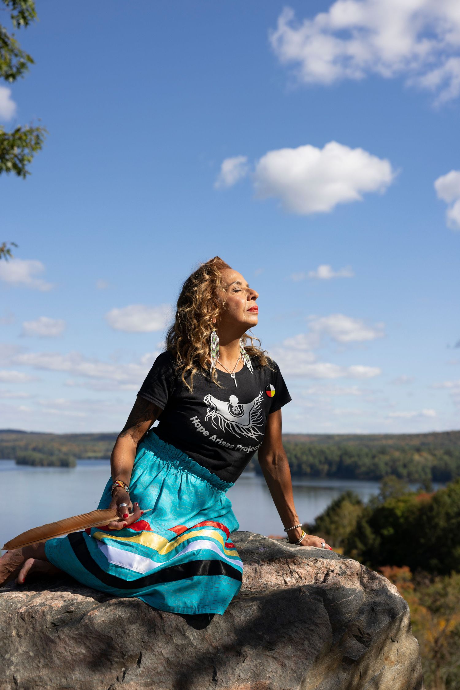 Person in black t-shirt and turquoise skirt sits peacefully on rocky overlook with lake and forest vista in background.