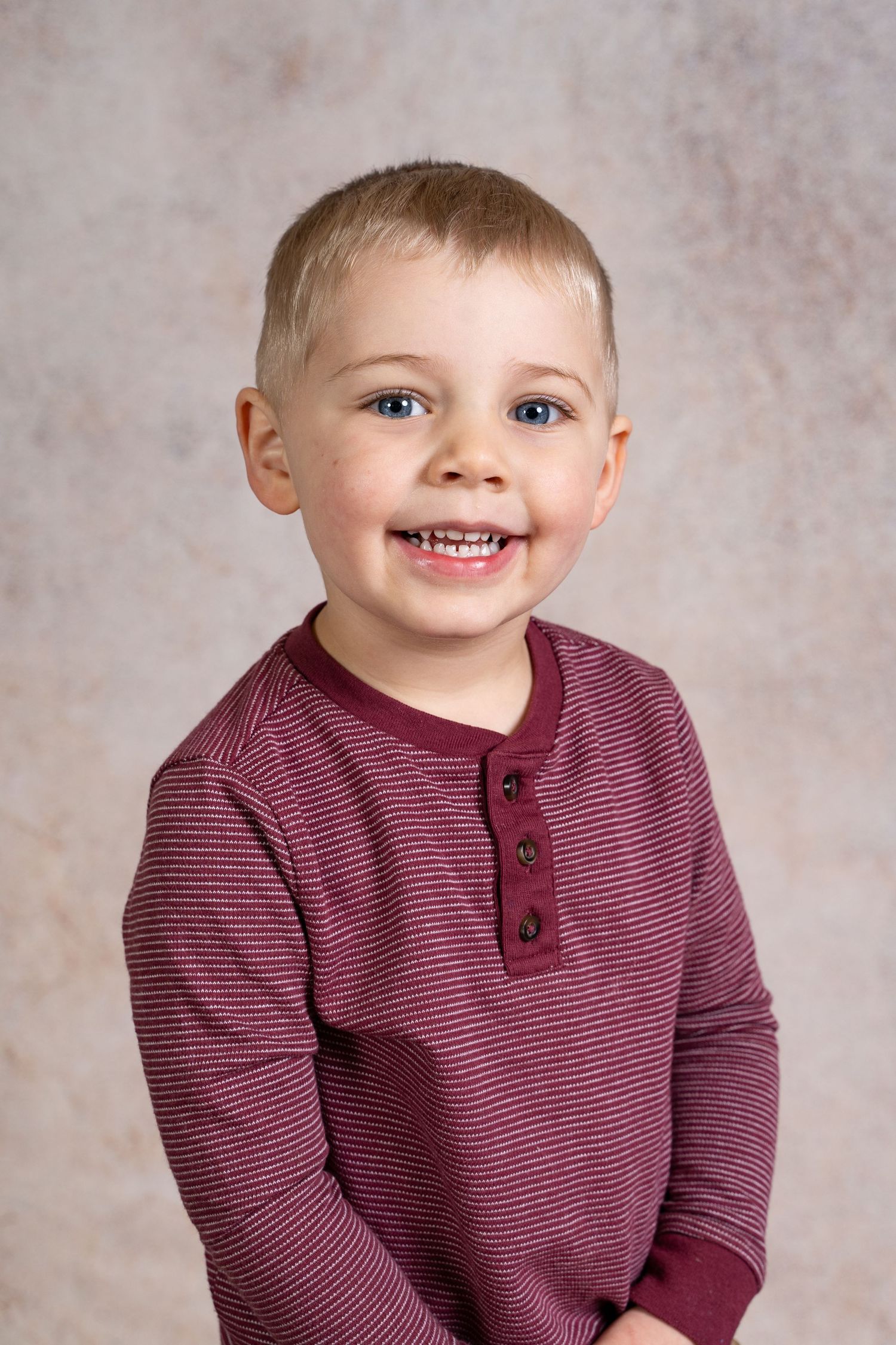A smiling child in a burgundy striped henley shirt poses against a textured gray backdrop.