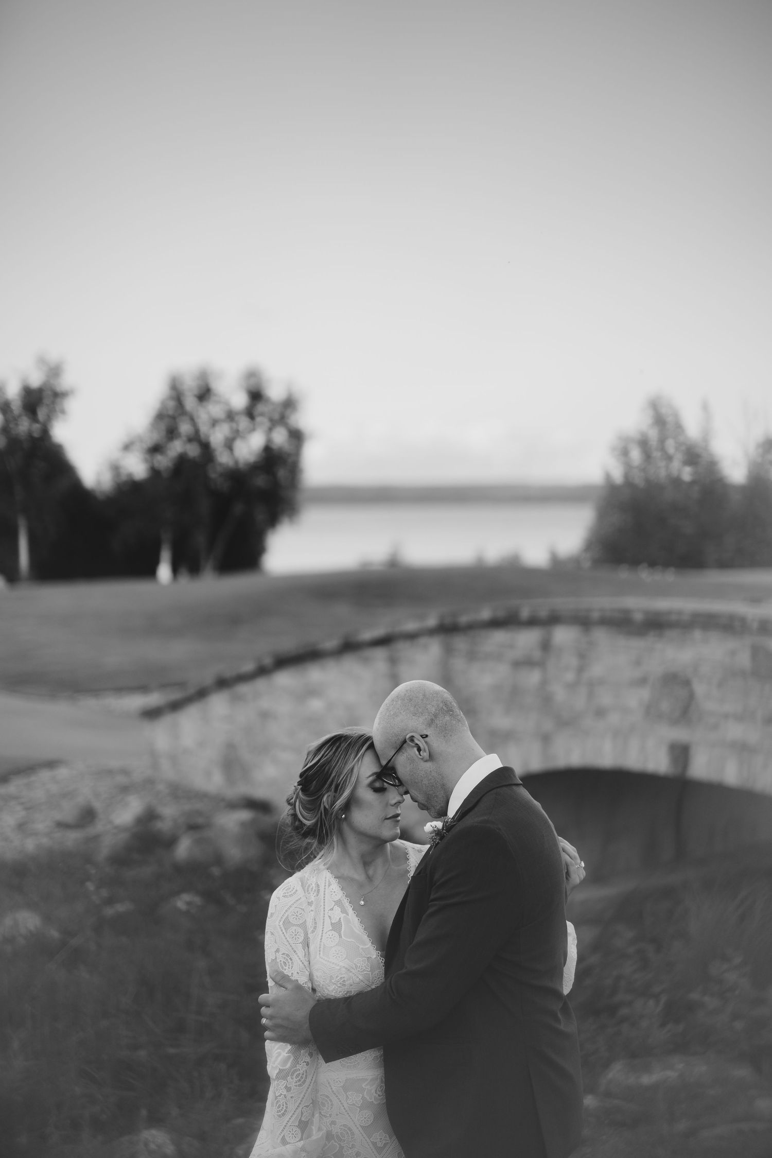 a black and white photo of a couple touch foreheads near a bridge in cobble beach