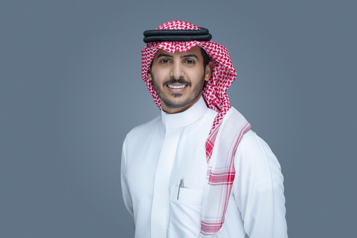 Young person in traditional Middle Eastern dress featuring white thobe and red and white checkered headscarf smiles at camera.