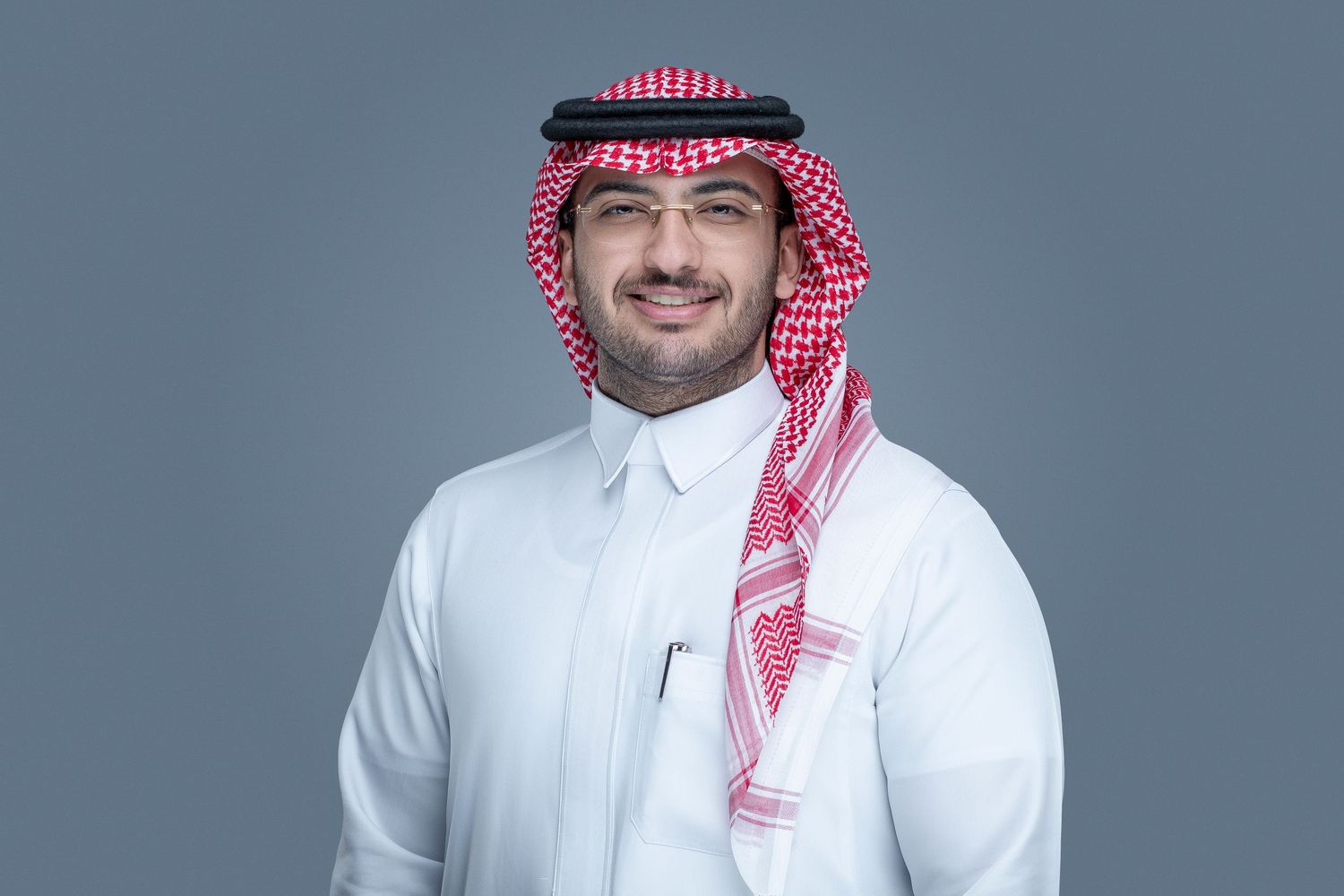 A person wearing traditional Middle Eastern dress including a white thobe and red and white checkered keffiyeh against a grey backdrop.