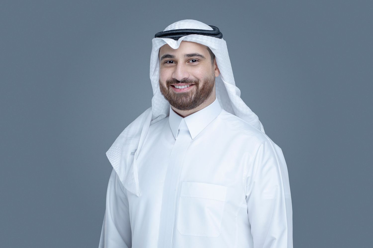 Man wearing traditional white Gulf Arab thobe and headdress standing against gray background with friendly expression.