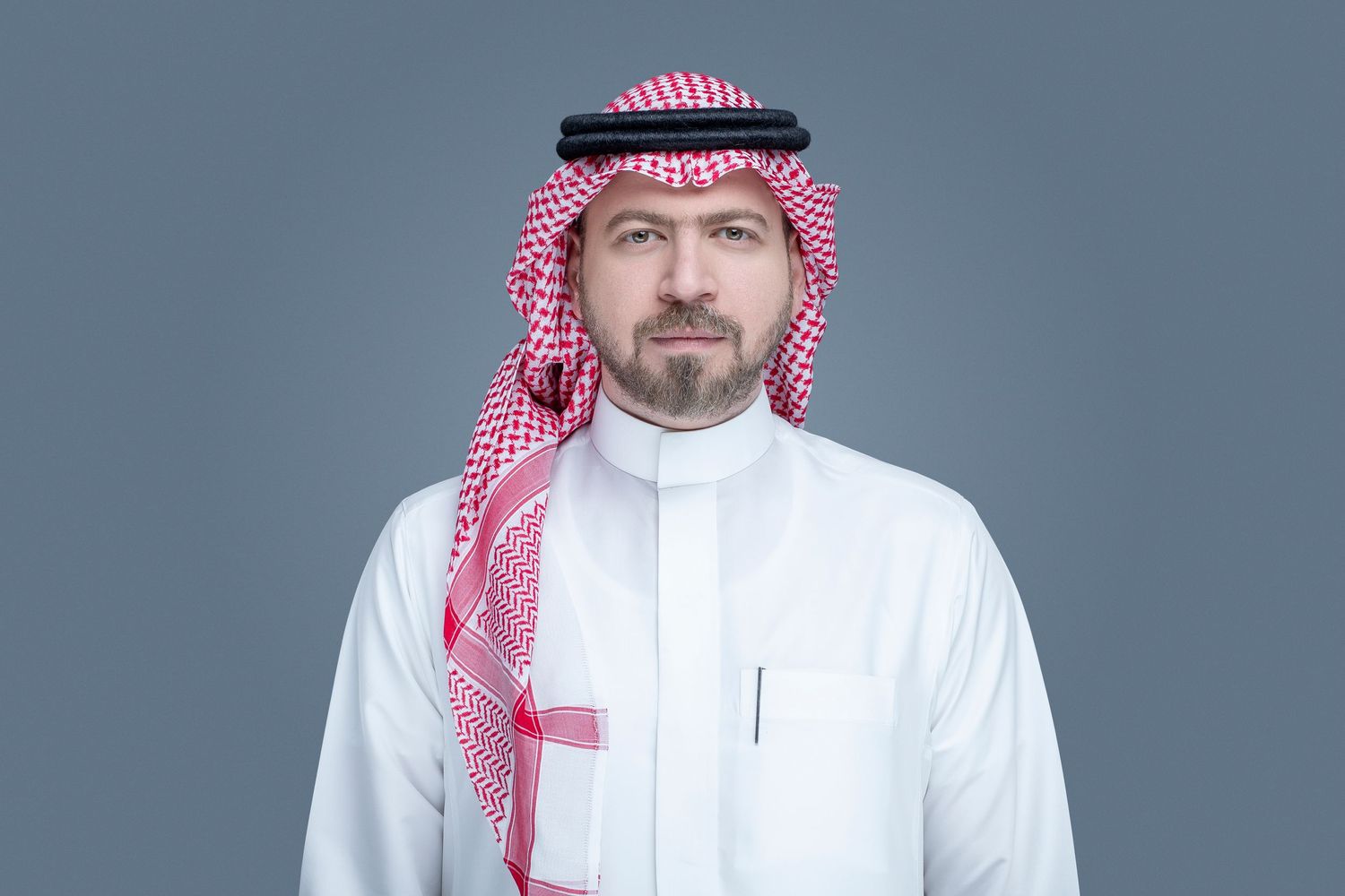 A person wearing traditional Middle Eastern attire including a white thobe and red and white checkered keffiyeh against a gray backdrop.