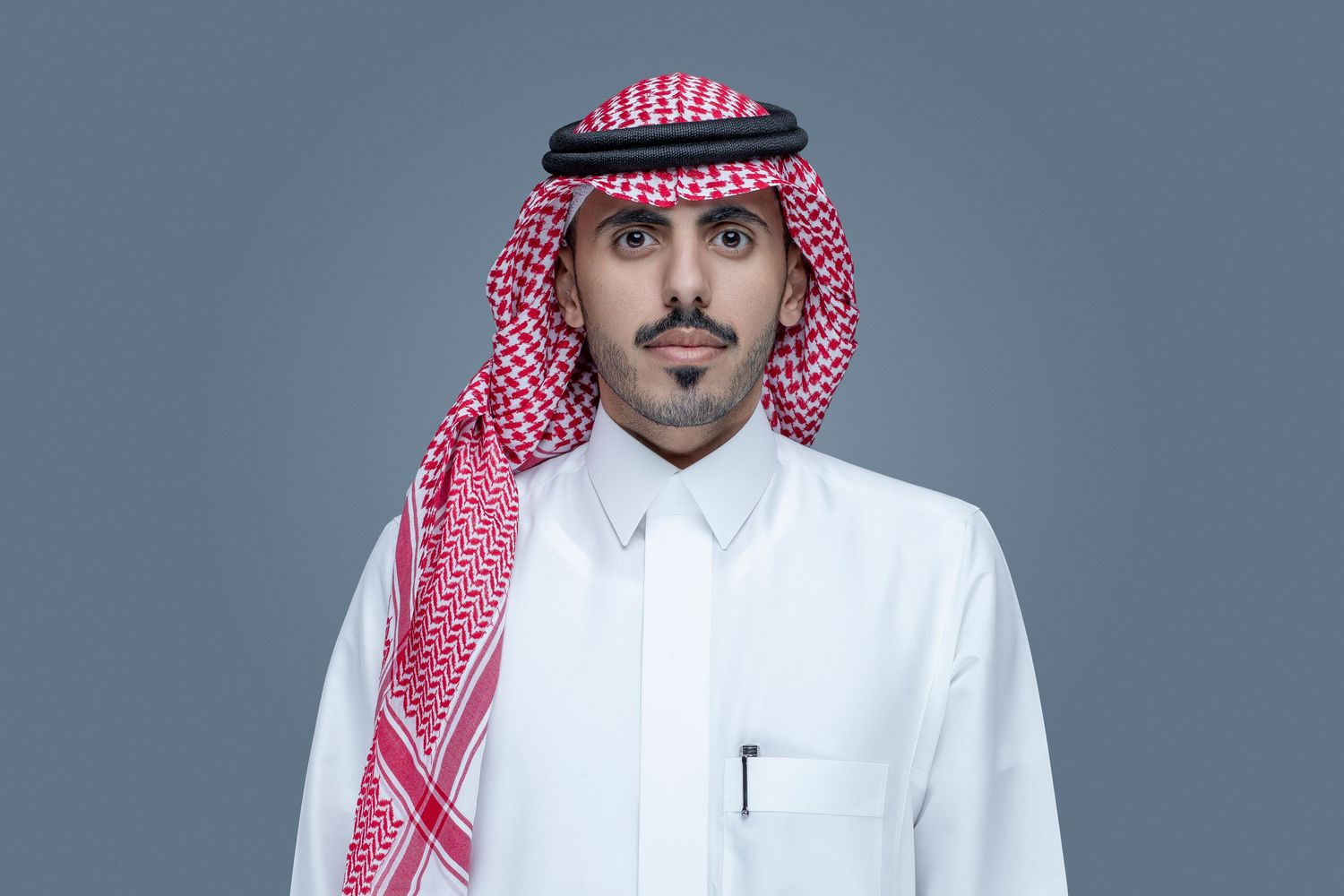 A person wearing a traditional red and white checkered keffiyeh headdress and crisp white thobe against a grey background.