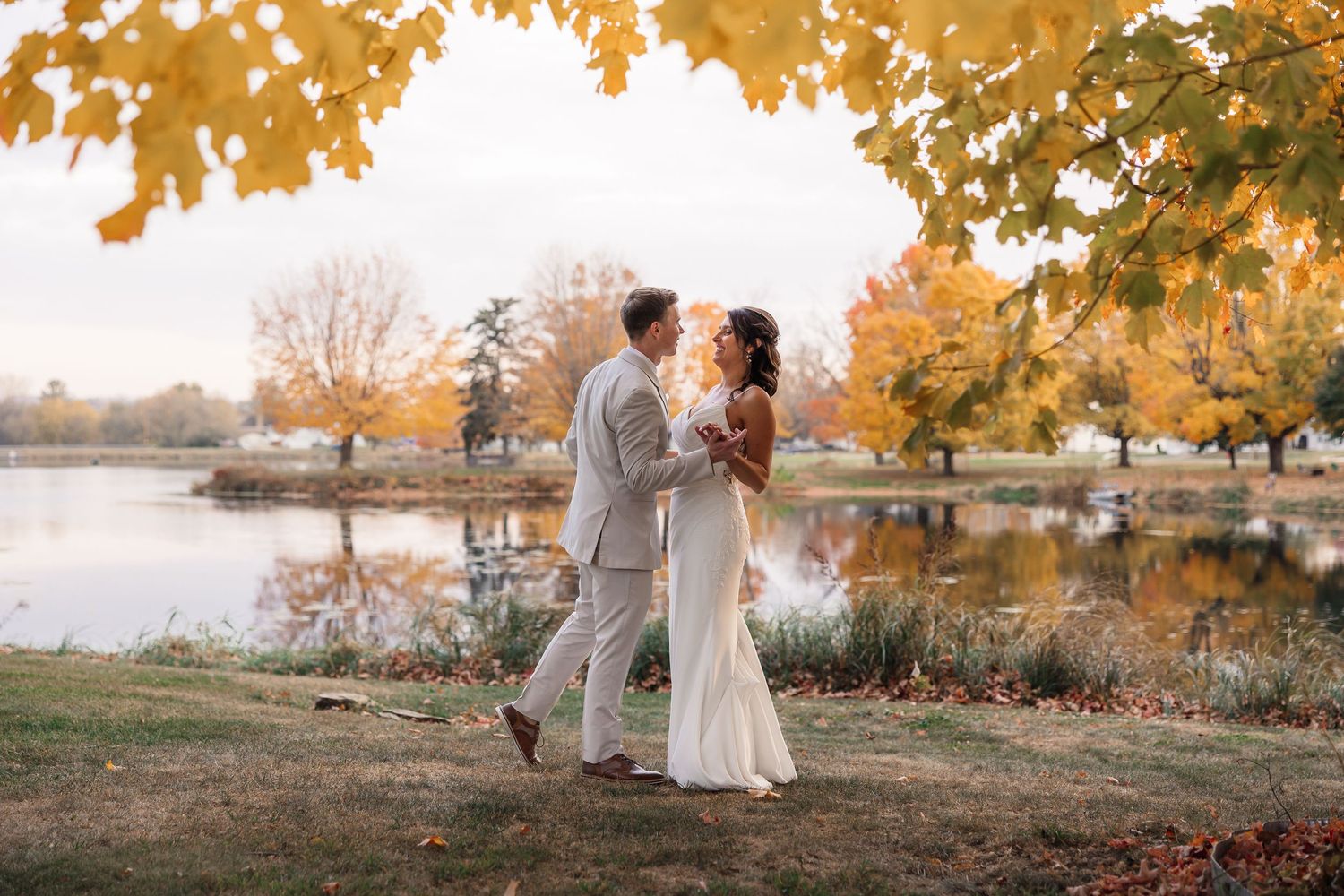 A couple embraces by a tranquil lake during autumn with golden leaves overhead.
