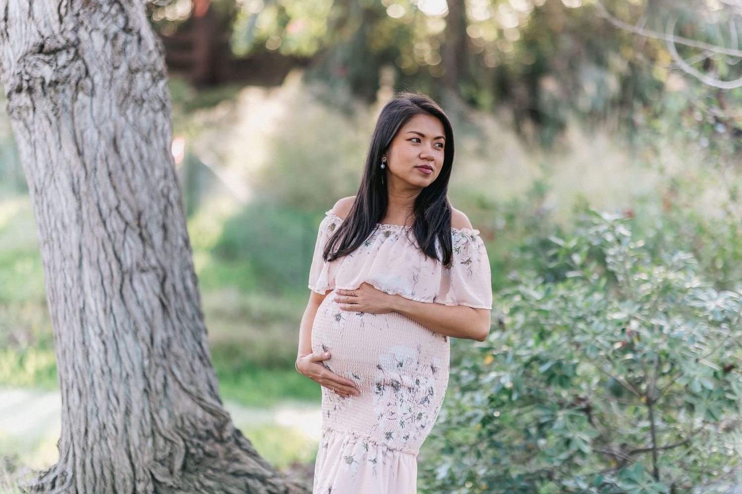 Maternity photo showing woman in white dress posing beside large tree trunk in garden setting.