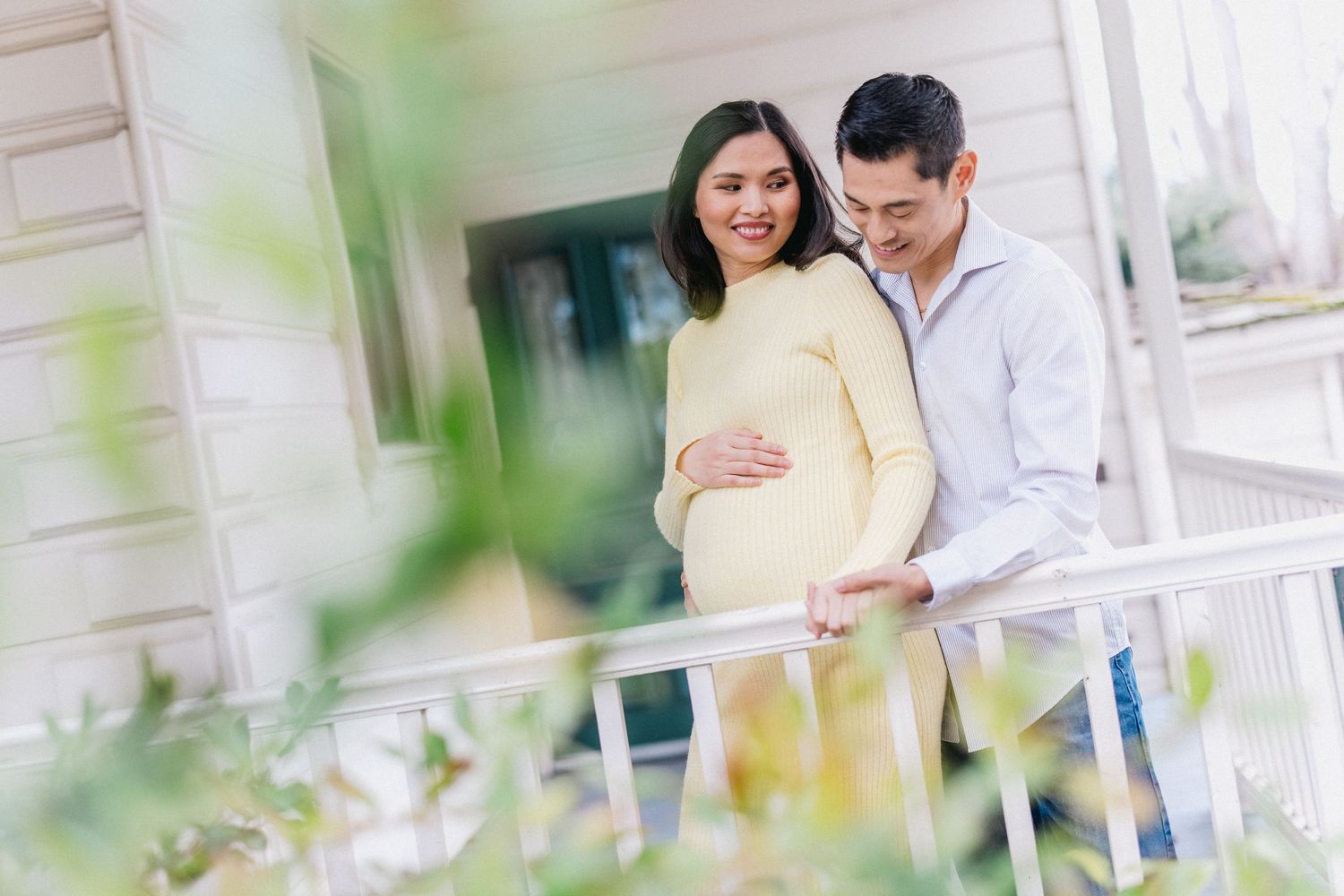 Intimate couple maternity photo on porch through greenery with soft focus.