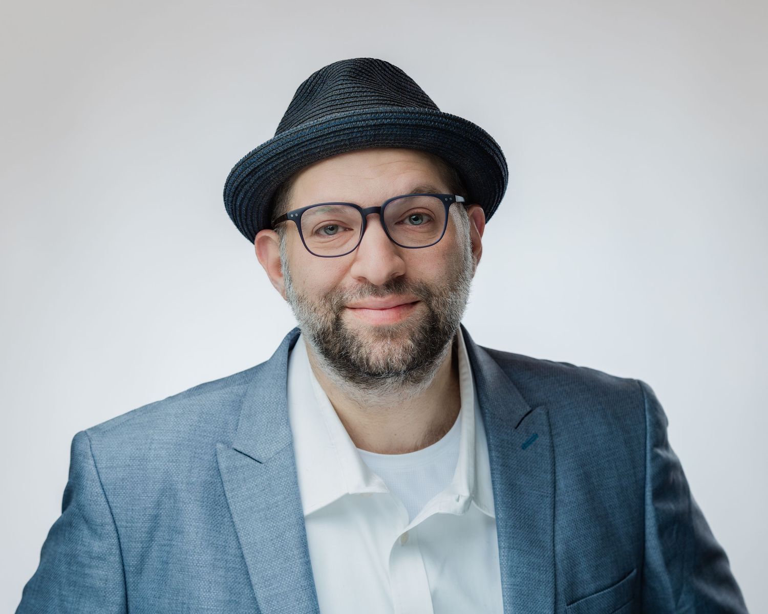 Professional headshot against white background of person wearing fedora, glasses and blue blazer with white shirt.