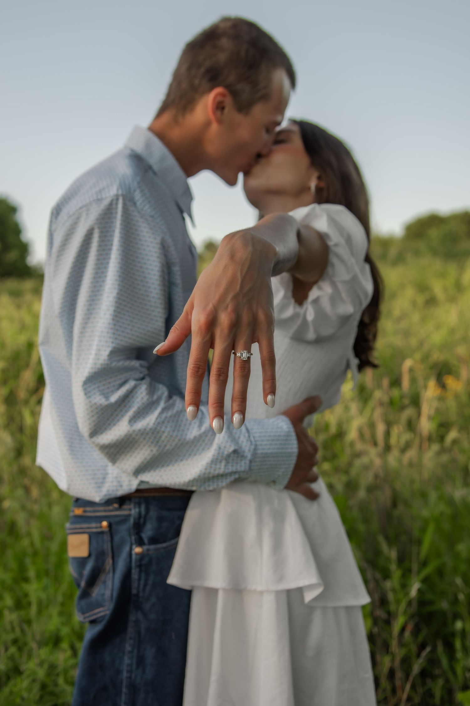 A couple in casual attire embraces each other in a grassy field during golden hour, highlighting the engagement ring.
