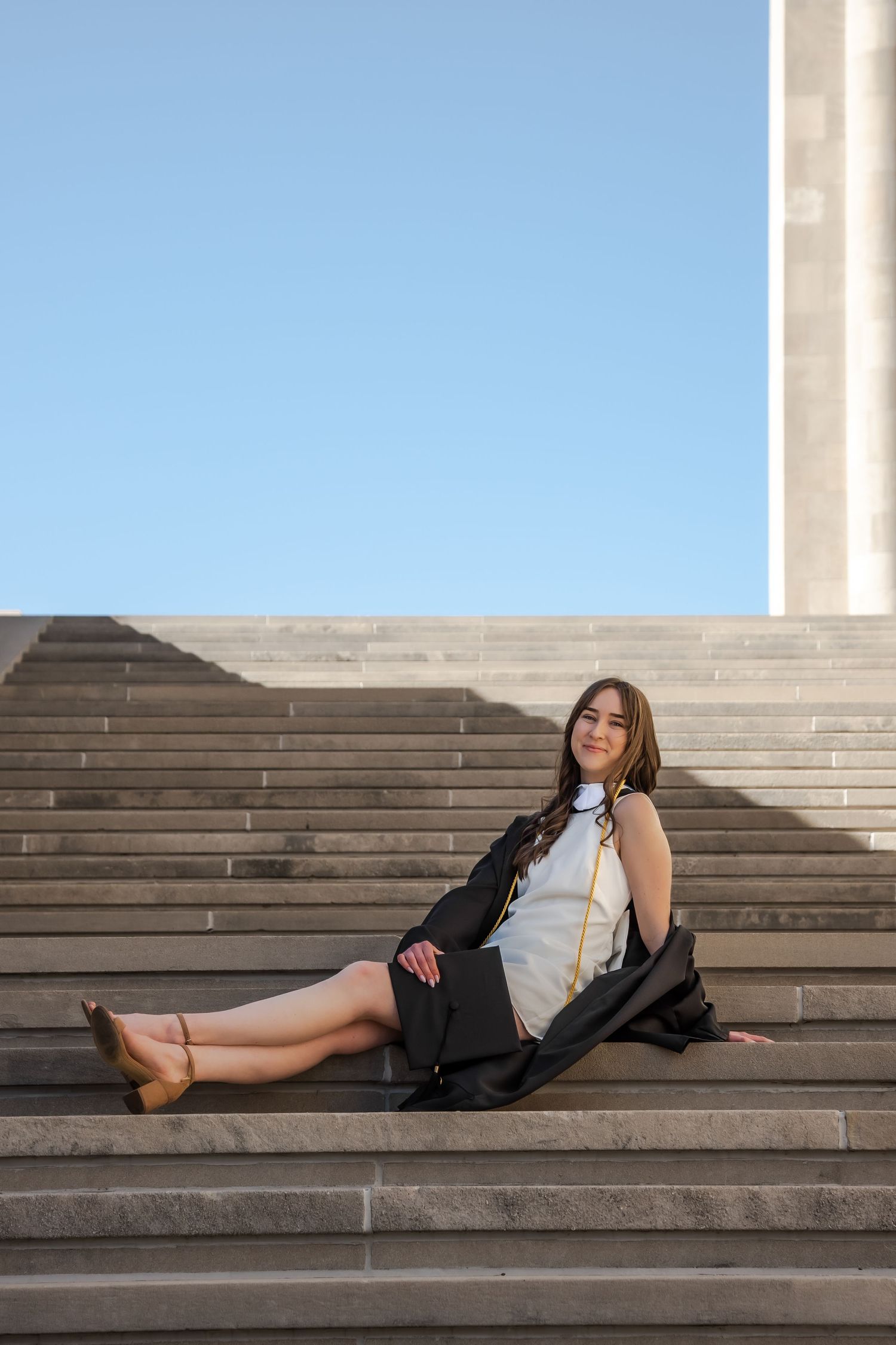 A person in a black blazer and white top reclines casually on concrete steps under bright sunlight.