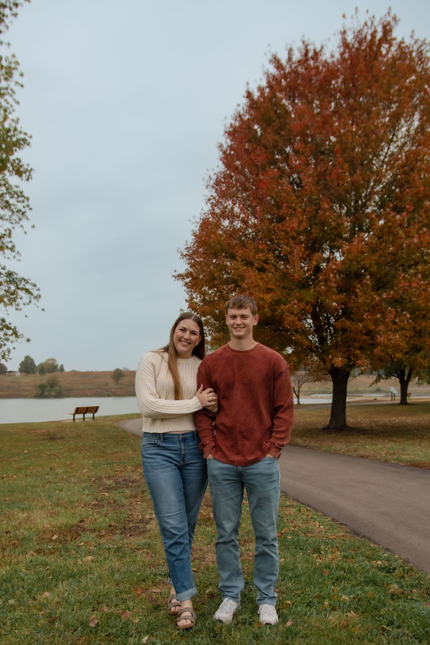 A couple in casual fall attire stands together on a winding path surrounded by autumn trees with orange foliage.
