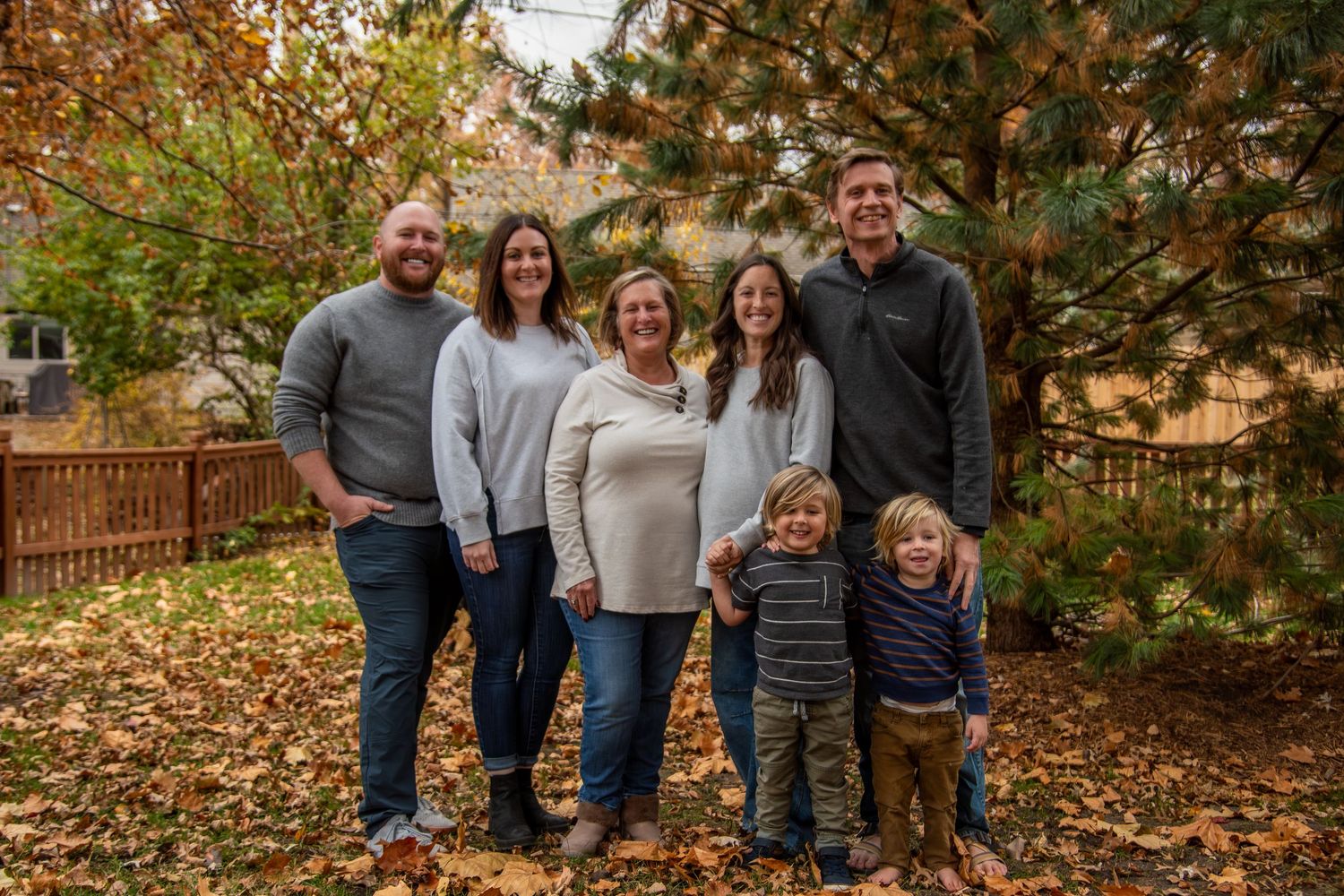 A group of people stand together outdoors surrounded by autumn foliage and fallen leaves in a backyard setting.