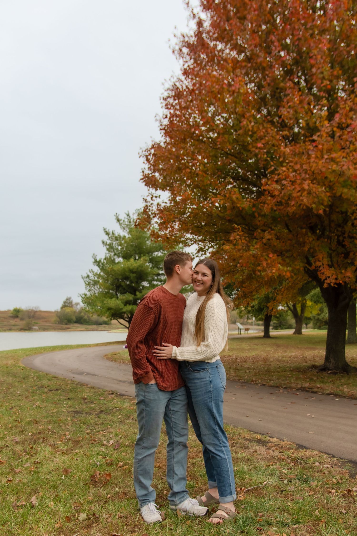 A couple embraces on a dirt path surrounded by autumn trees with vibrant orange foliage on a cloudy day.