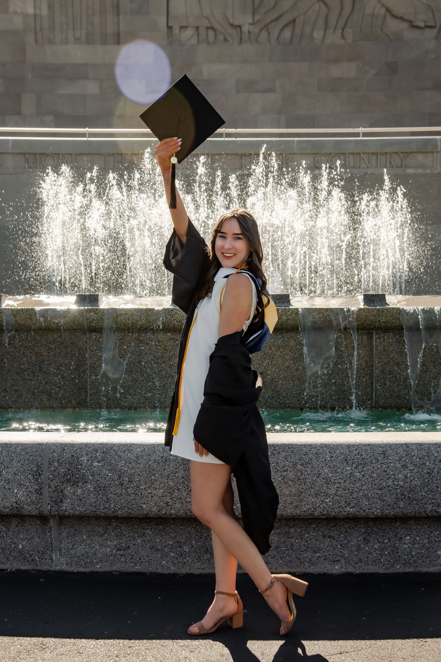 Graduate in black gown and white dress posing playfully by decorative water fountain.