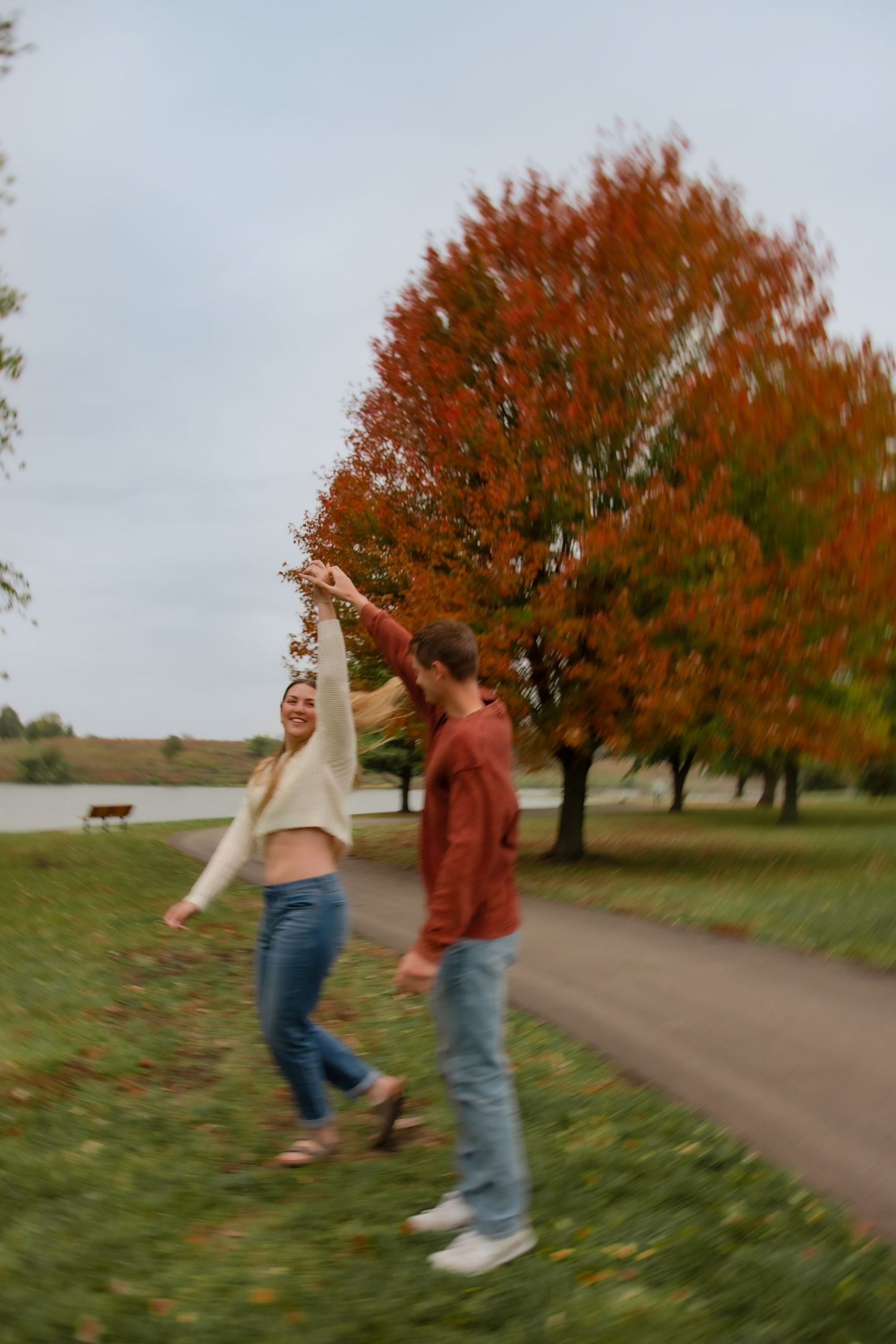 Two people dance and spin joyfully on a path surrounded by autumn trees with vibrant red and orange foliage.