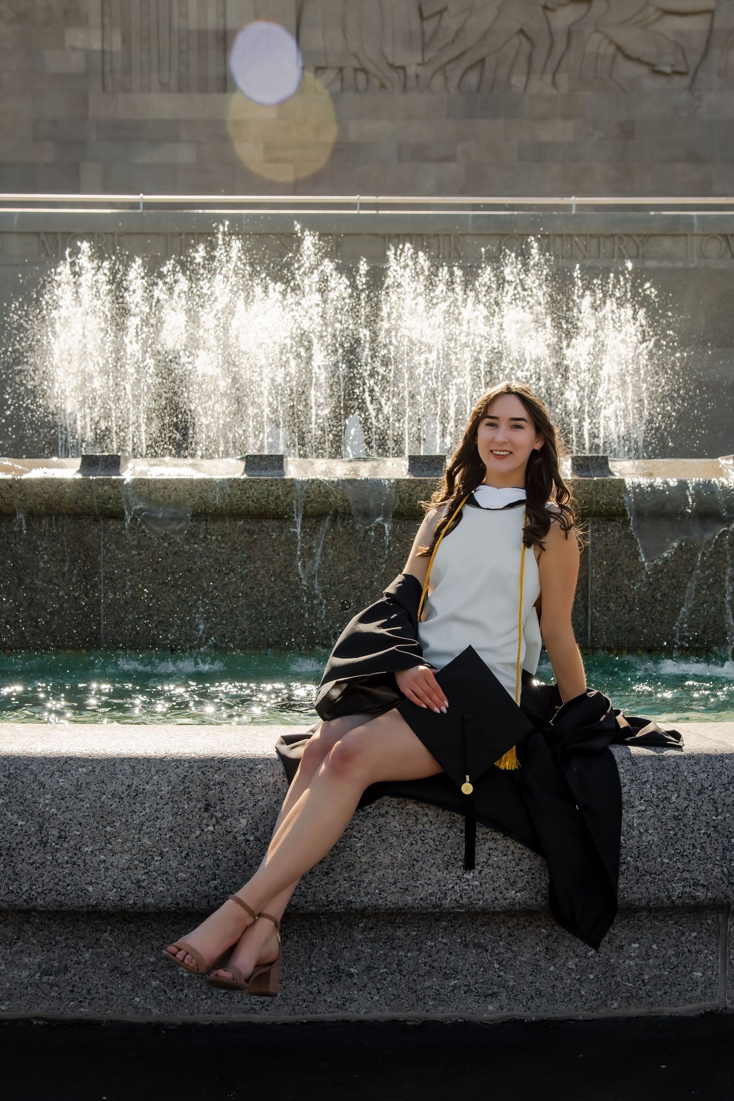 Graduate in academic regalia sitting casually by outdoor fountain during photo session.