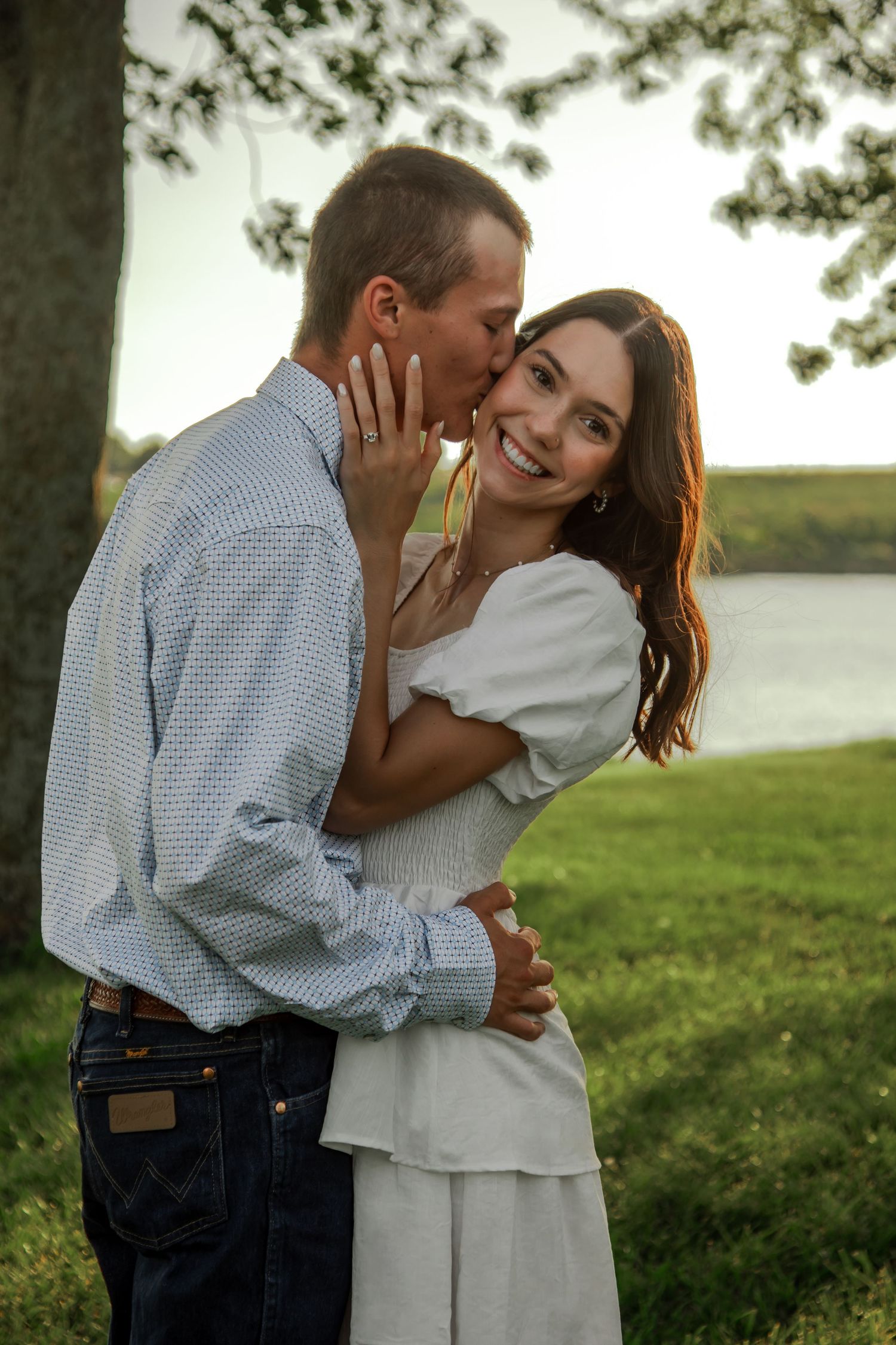 A romantic couple embraces on a grassy hillside near a lake at sunset.