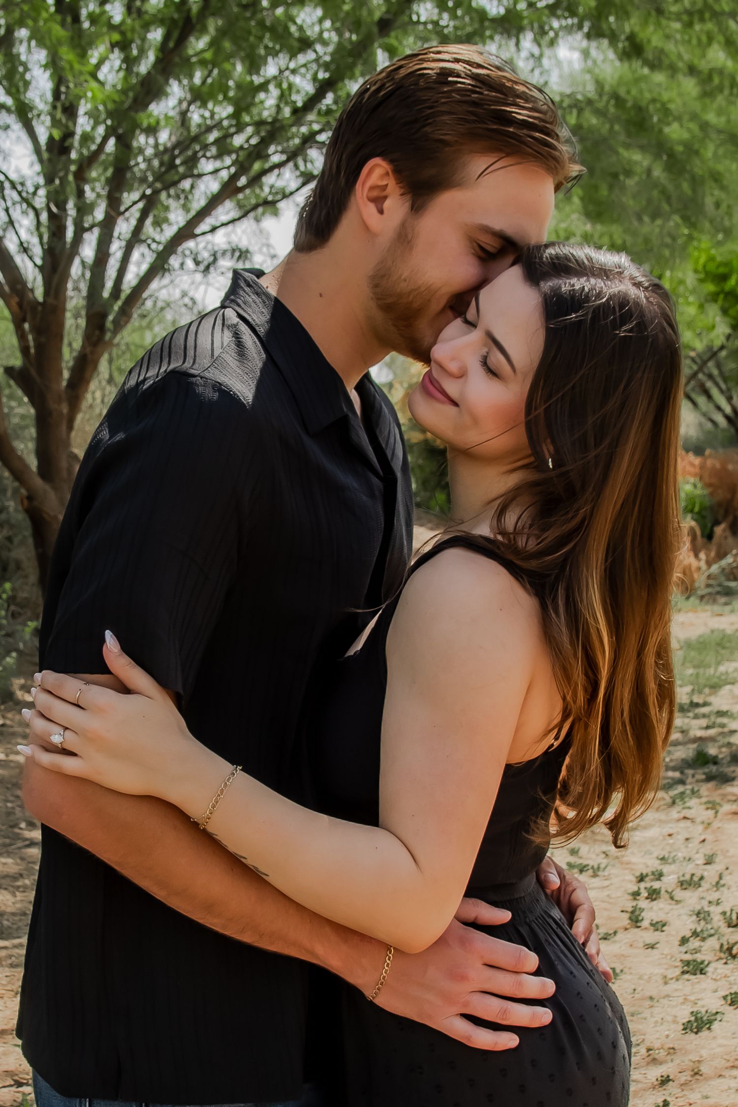 A couple in black attire share an intimate embrace while standing outdoors among trees and desert landscape.