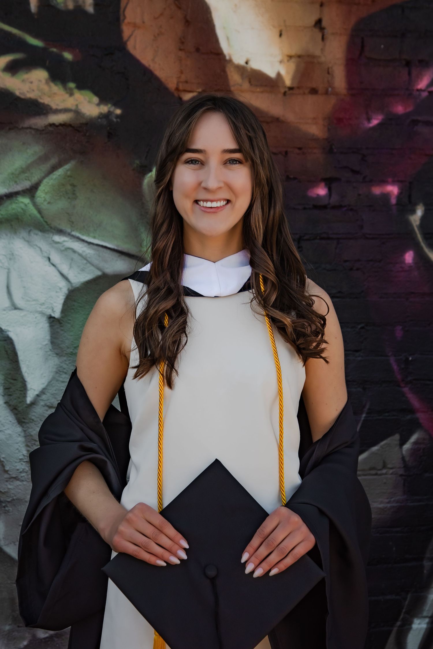 Smiling young woman in black graduation gown and gold honor cord holding cap in front of colorful mural.