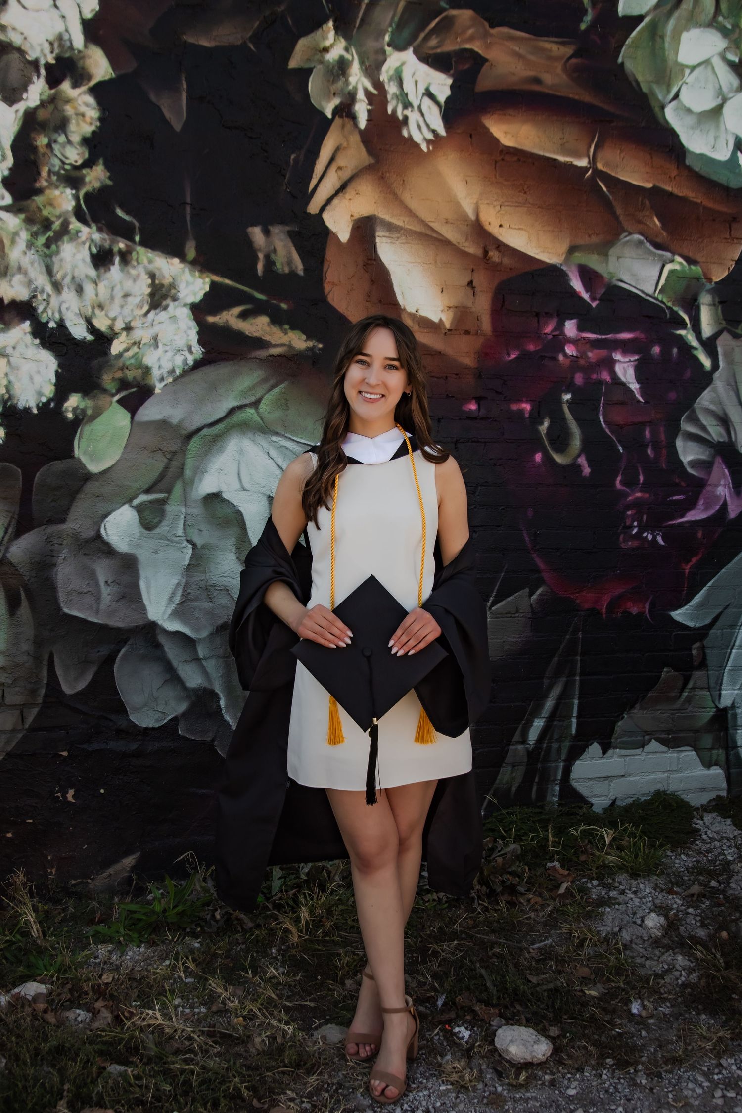 Graduate in white dress and black gown poses against floral mural backdrop.