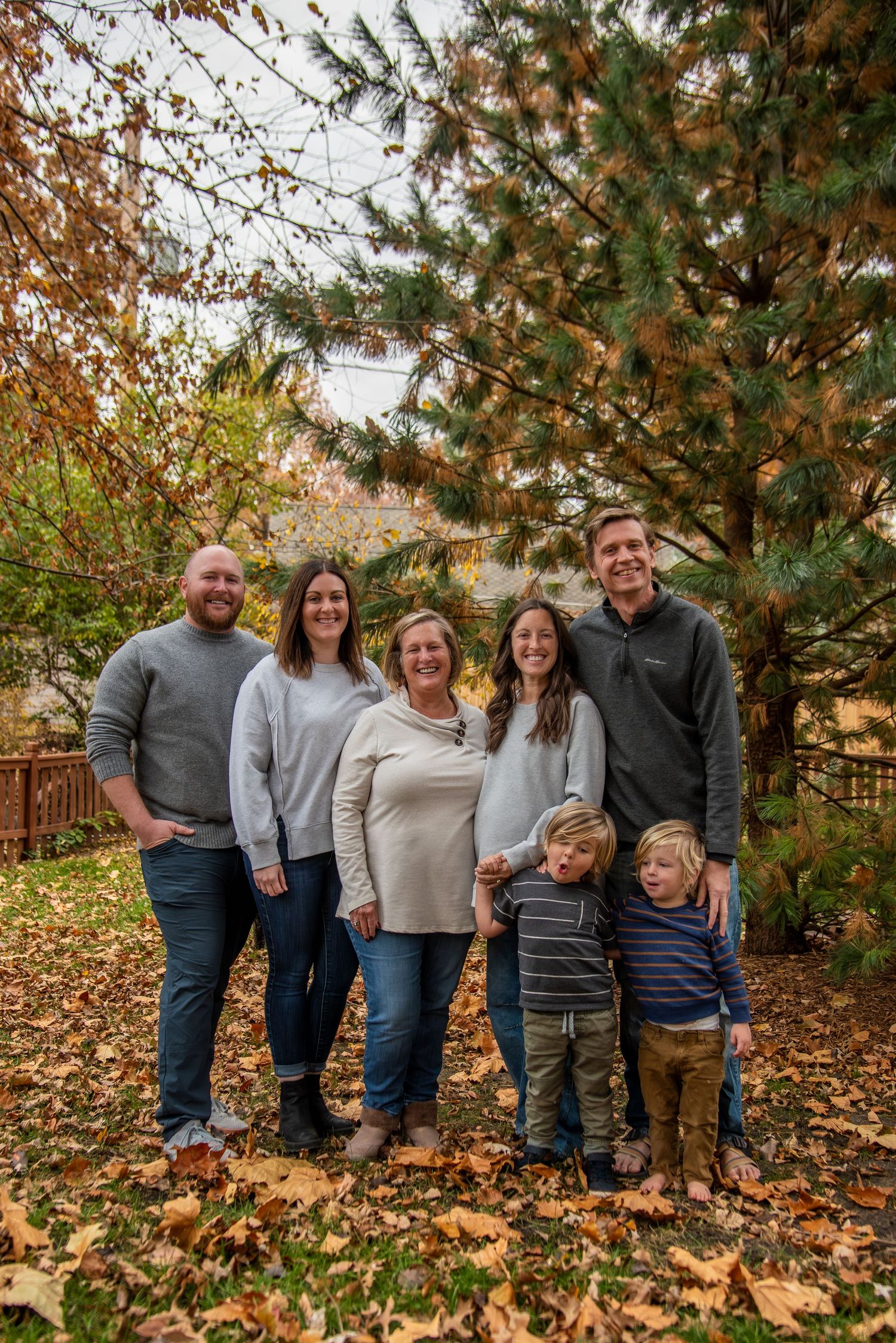 A family poses together outdoors on an autumn day with fallen leaves and evergreen trees in the background.