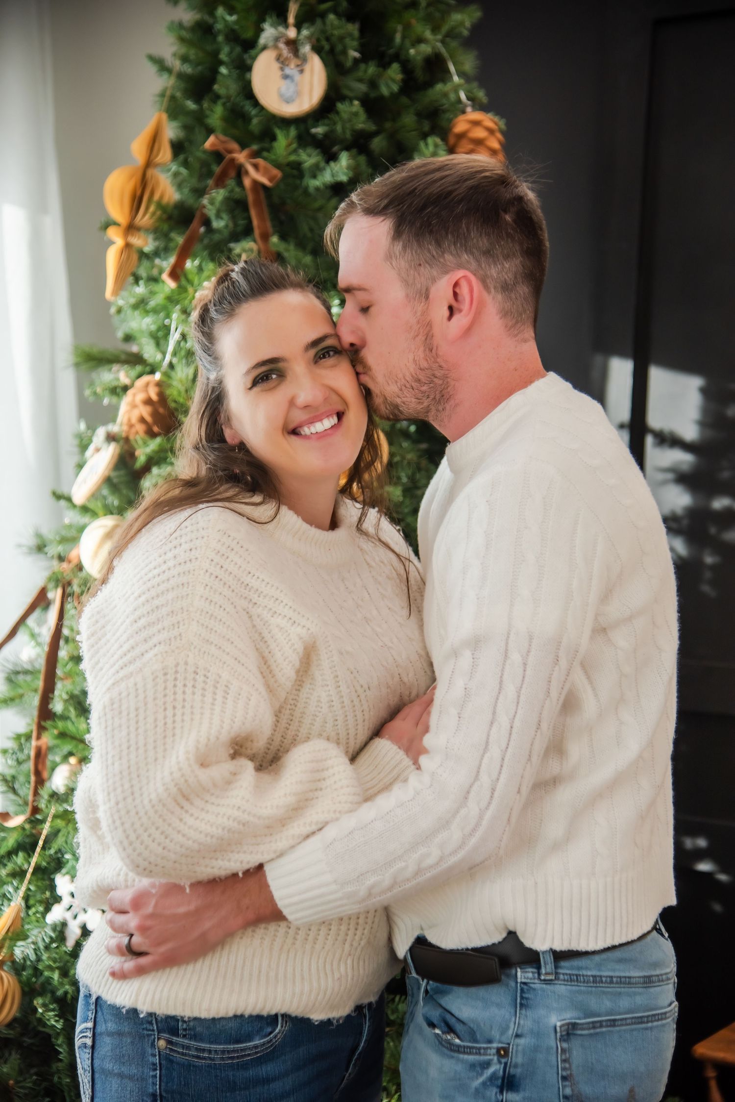 Couple in matching cream cable knit sweaters share intimate holiday moments by a decorated Christmas tree.