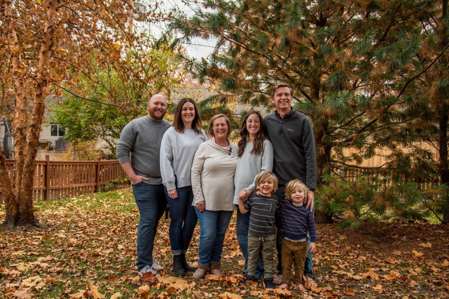 Family members gather outdoors for a casual photo shoot among scattered fall foliage and wooden fencing.