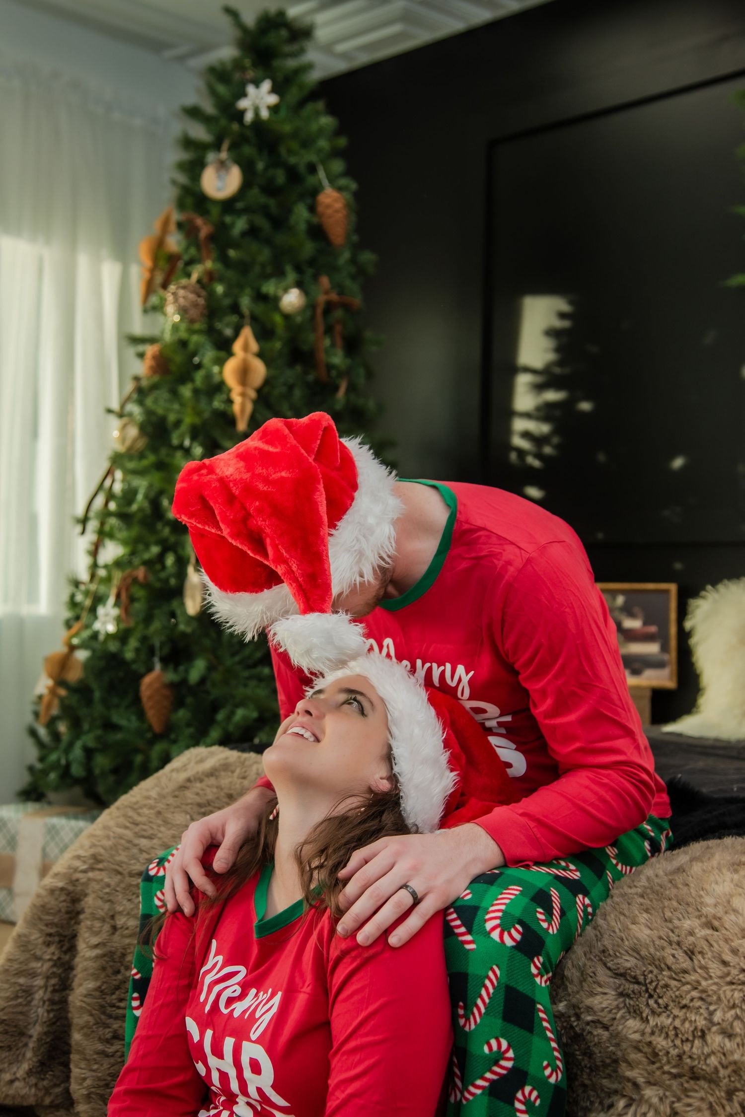 Playful holiday scene with couple in matching Christmas pajamas and Santa hats lounging on brown couch.