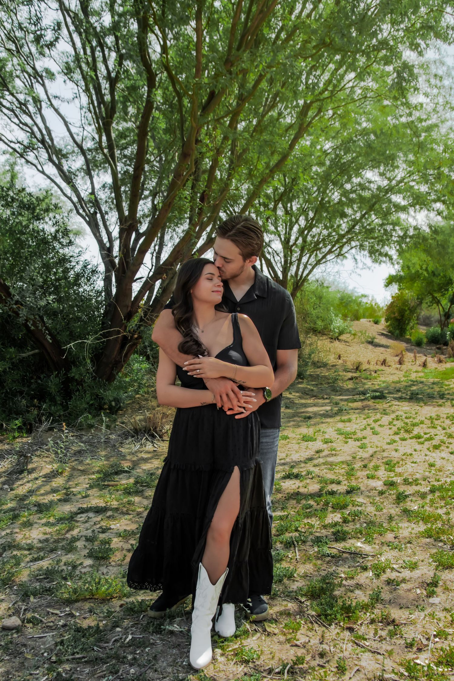 A couple in coordinated black and white outfits share intimate romantic moments under green trees in a desert landscape.